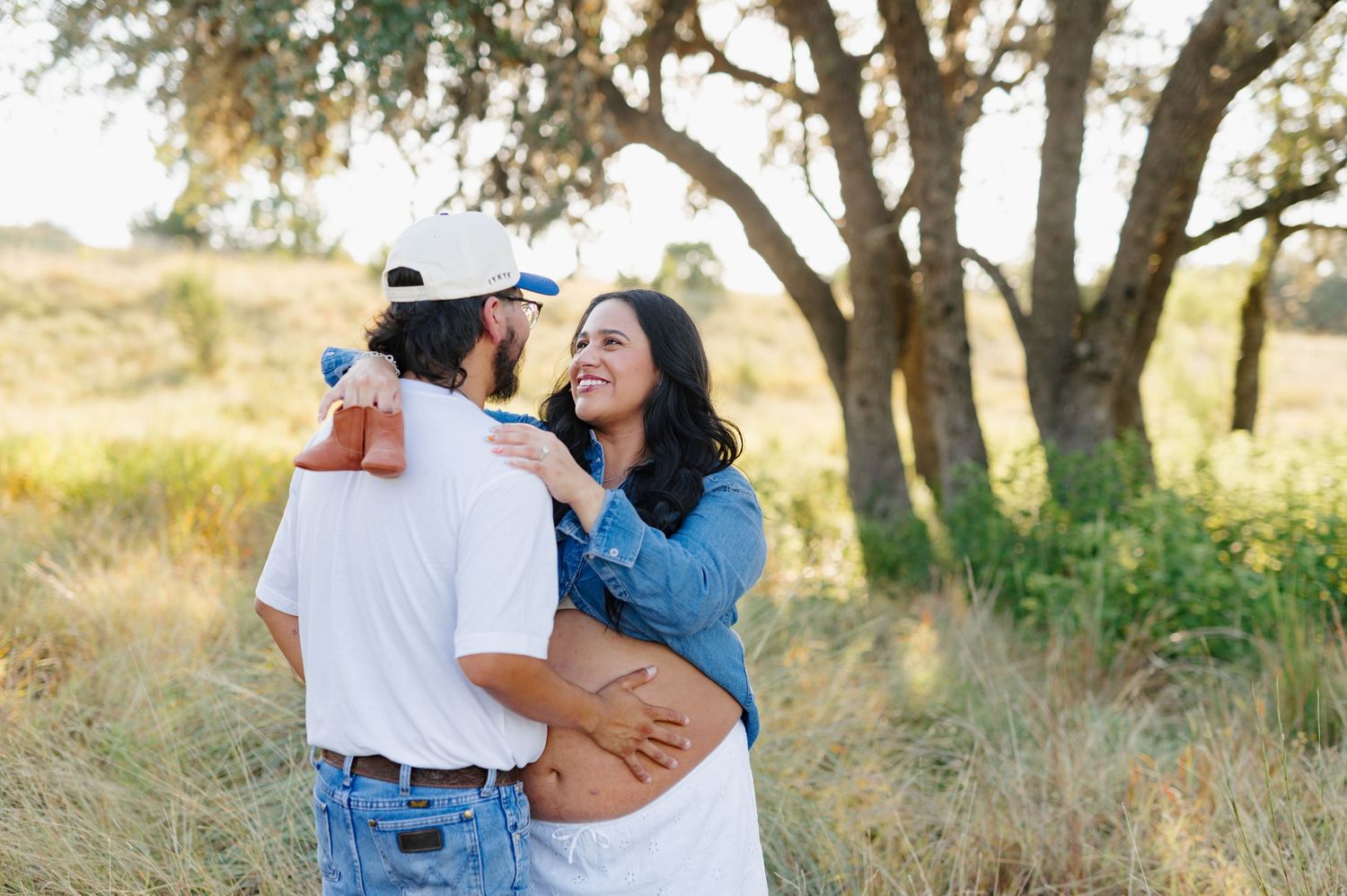 A tender moment captured during an outdoor maternity photoshoot among trees at golden hour.