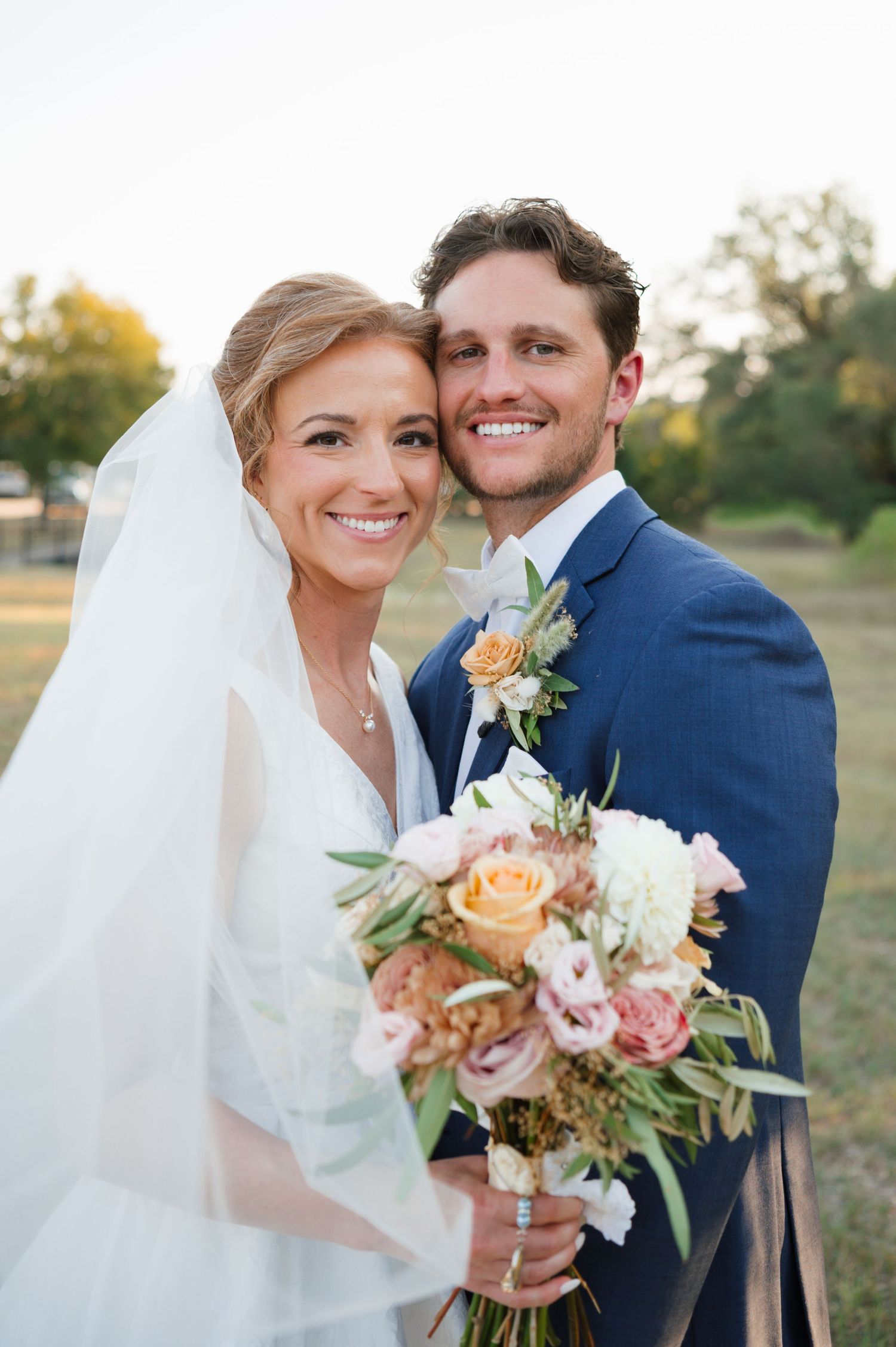 A happy couple in wedding attire poses outdoors with a colorful bouquet of flowers at sunset.