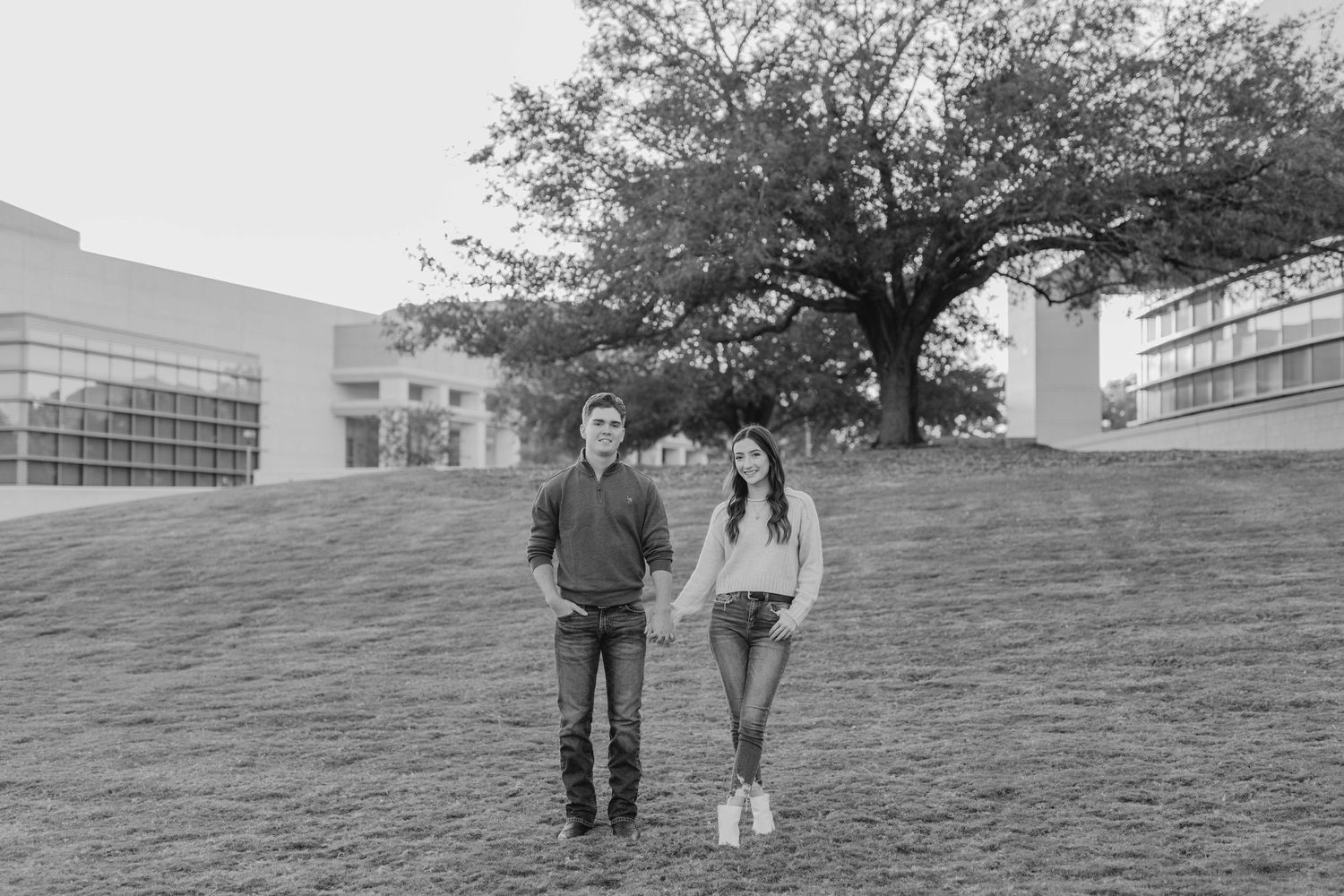 A couple stands together on a grassy hillside in black and white with modern buildings behind them.