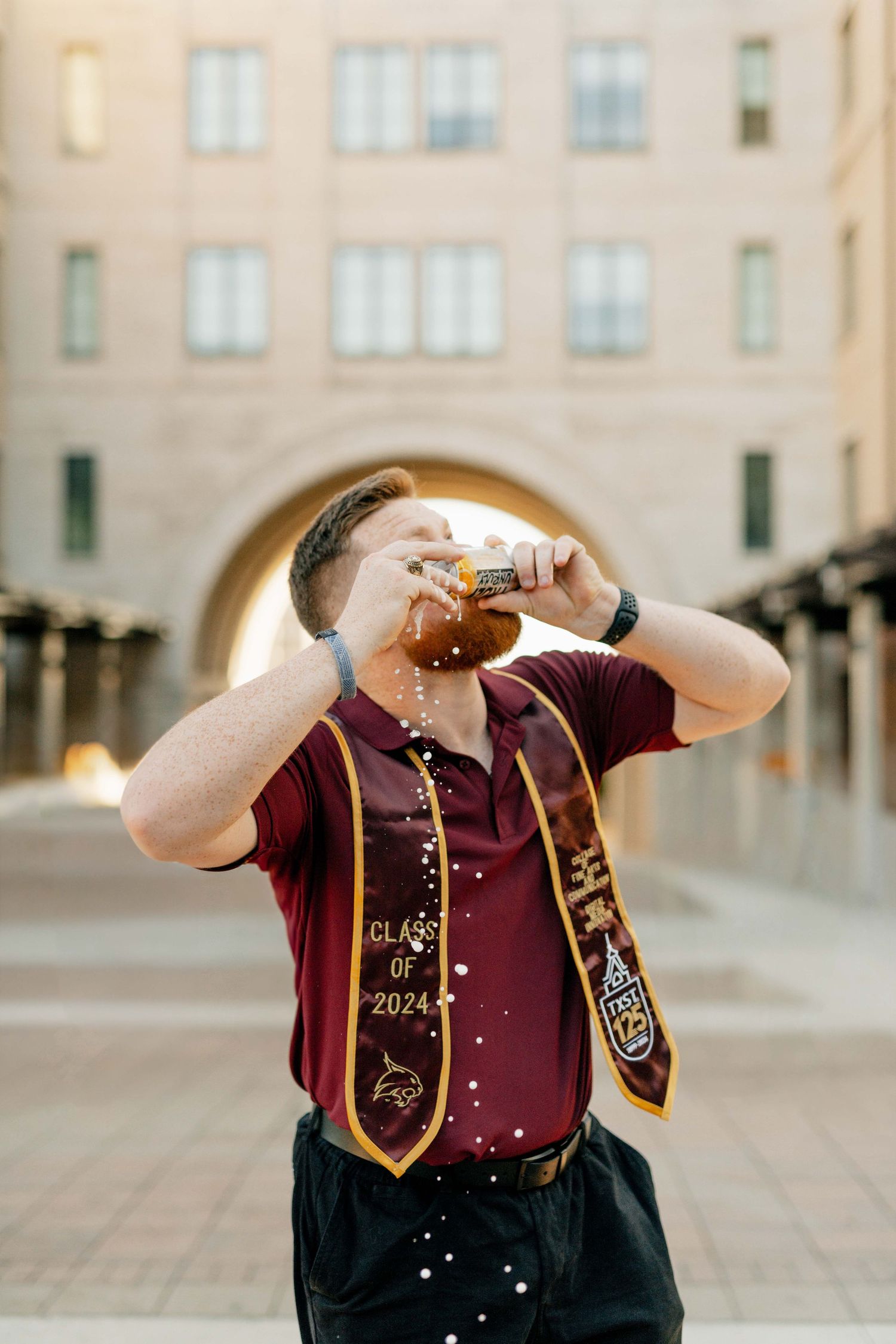 Graduate celebrates by popping champagne in front of campus building archway.