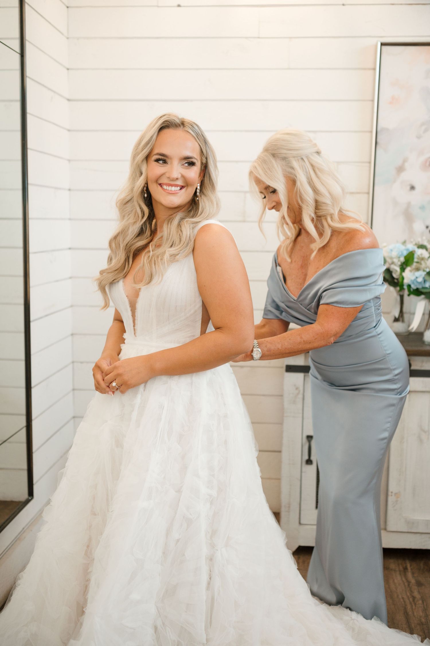 A bride in a white wedding dress is being embraced by a woman in a gray dress.