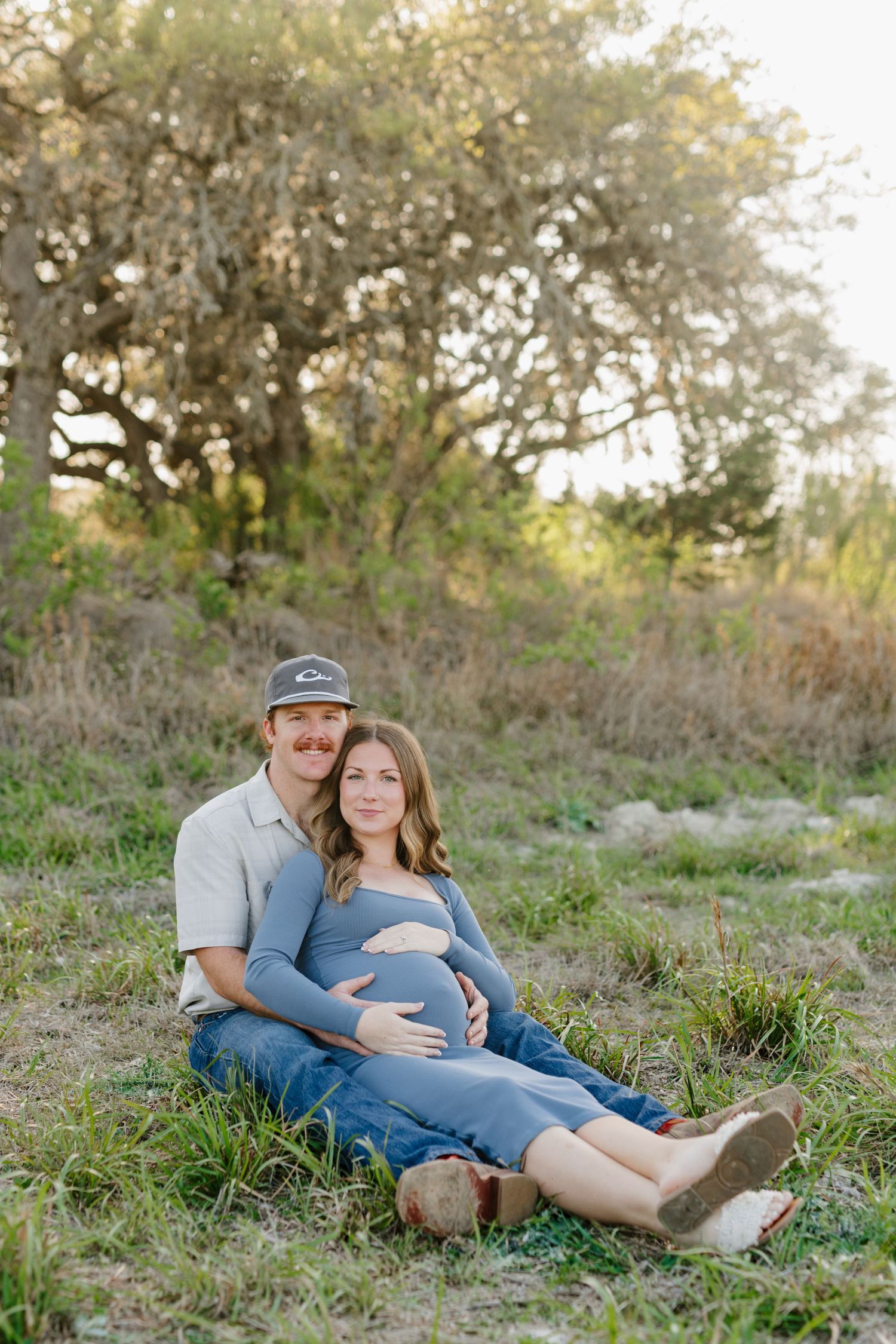 A couple relaxes on grassy ground during sunset maternity photoshoot in a natural outdoor setting.