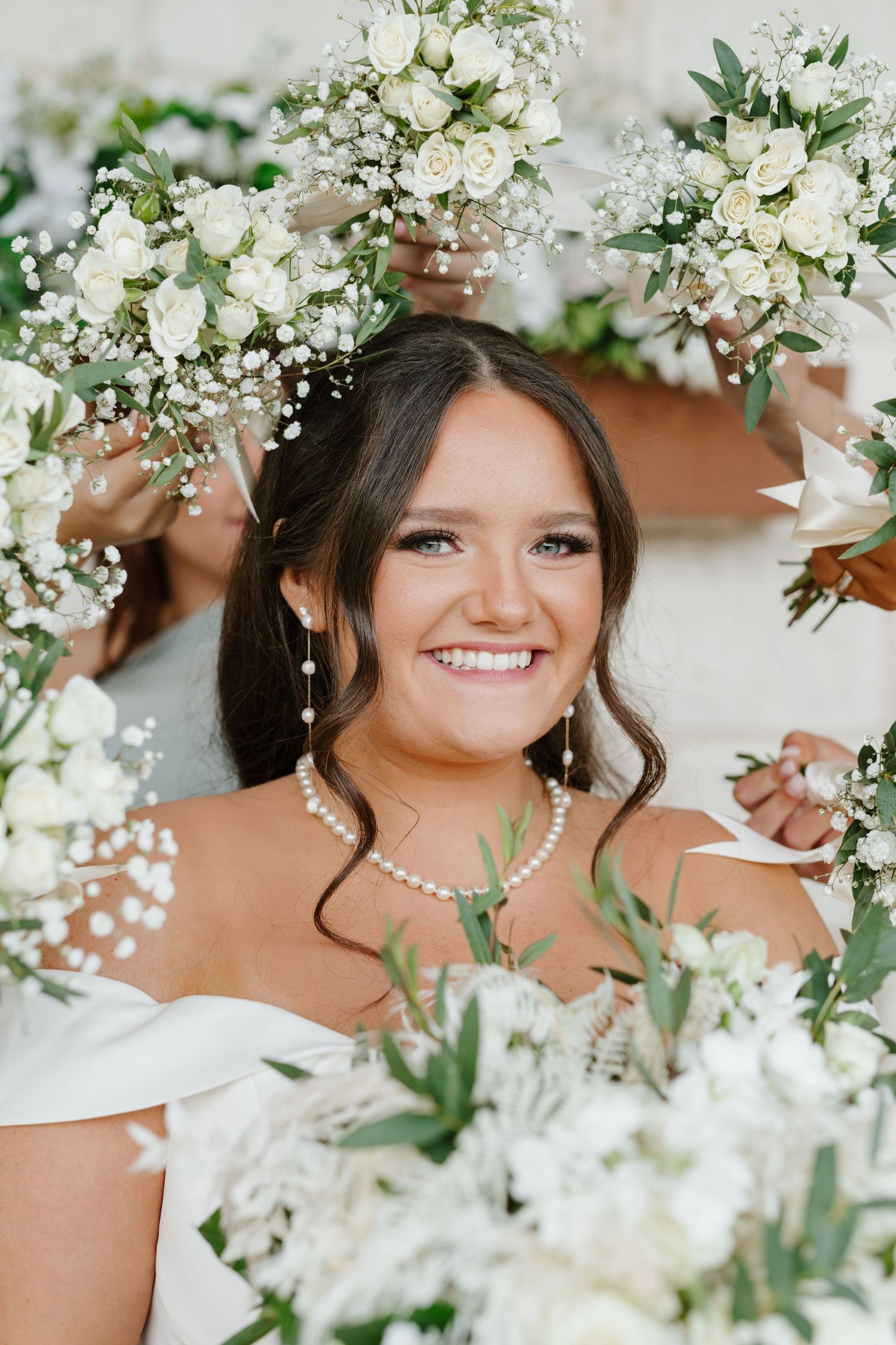 A radiant bride surrounded by white floral bouquets and baby's breath wearing pearl jewelry and an off-shoulder dress.