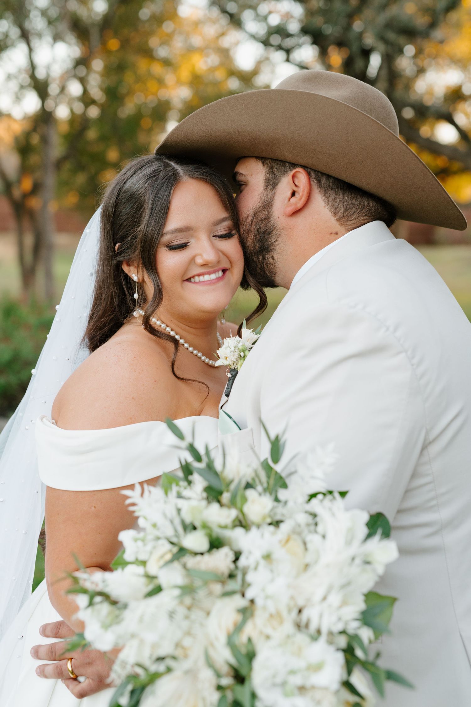 A loving couple celebrates their wedding day outdoors with white floral bouquet and western style attire.