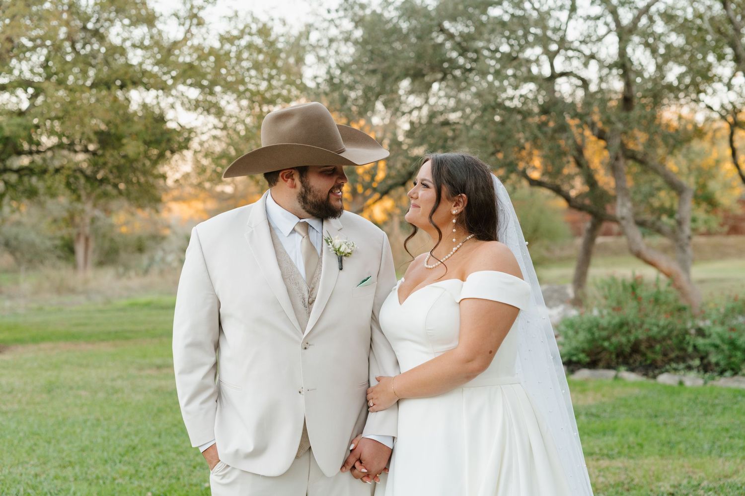 A couple in wedding attire holds hands in a rustic outdoor setting with trees and sunlit grass in the background.