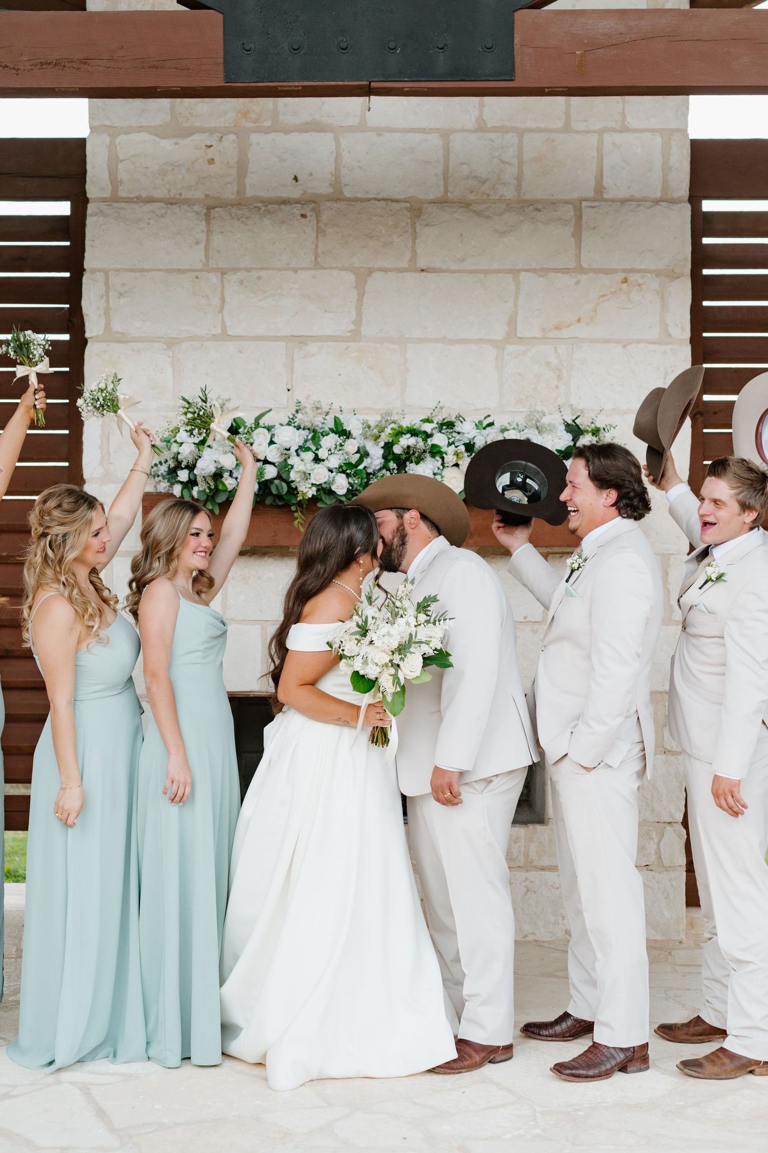 Wedding party in mint green bridesmaid dresses and beige suits pose together under stone archway with floral decor.