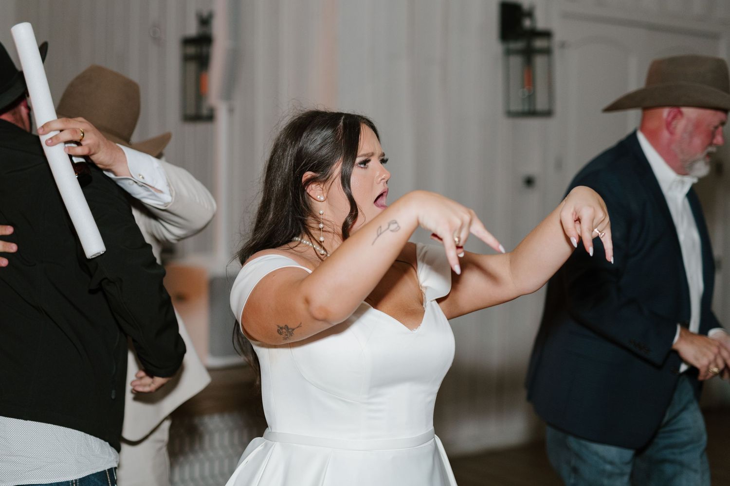 Wedding guests dancing enthusiastically on the dance floor at an indoor reception venue with wall sconces.