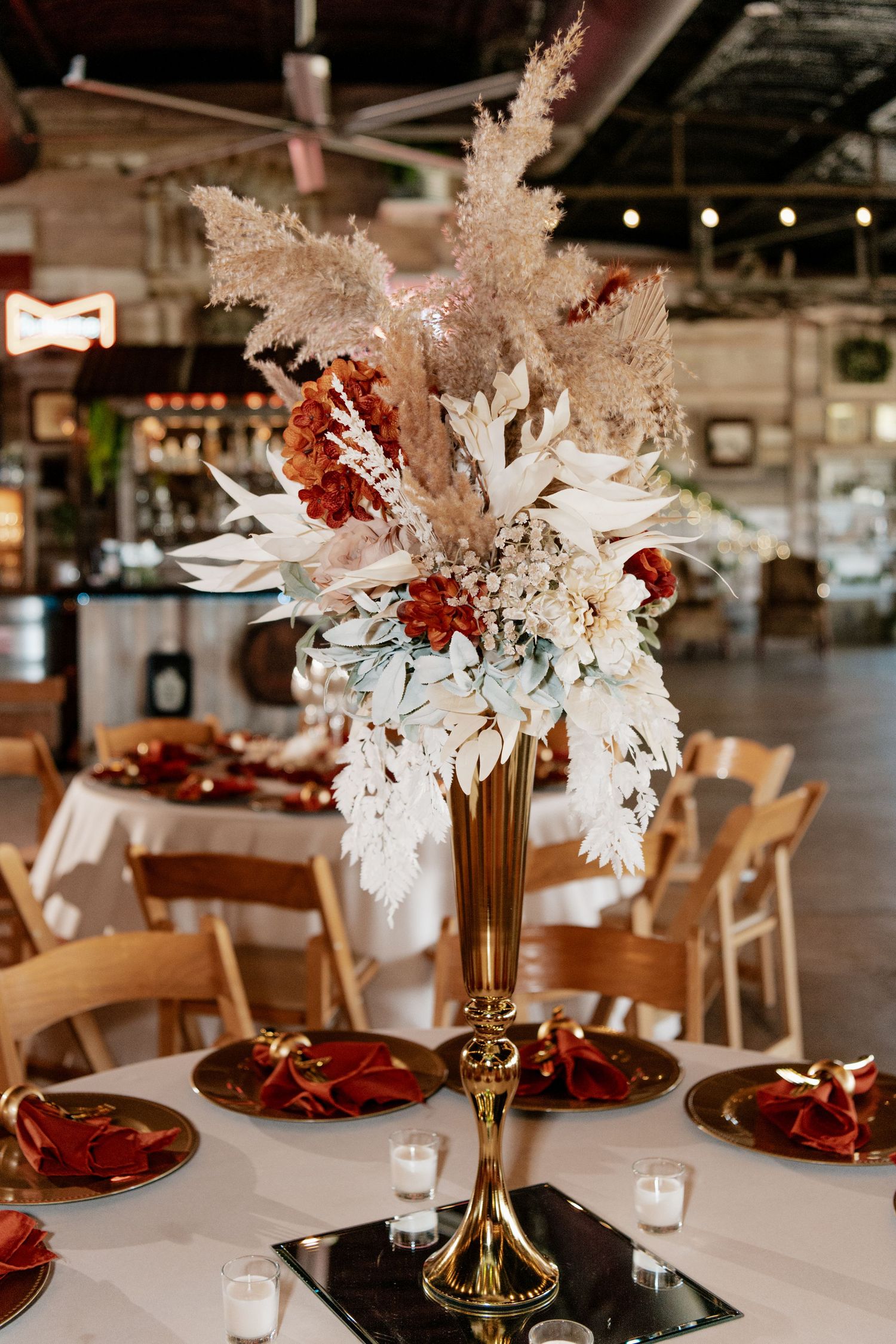 Elegant wedding reception centerpiece with dried pampas grass and red accents in a gold stand on white tablecloth.