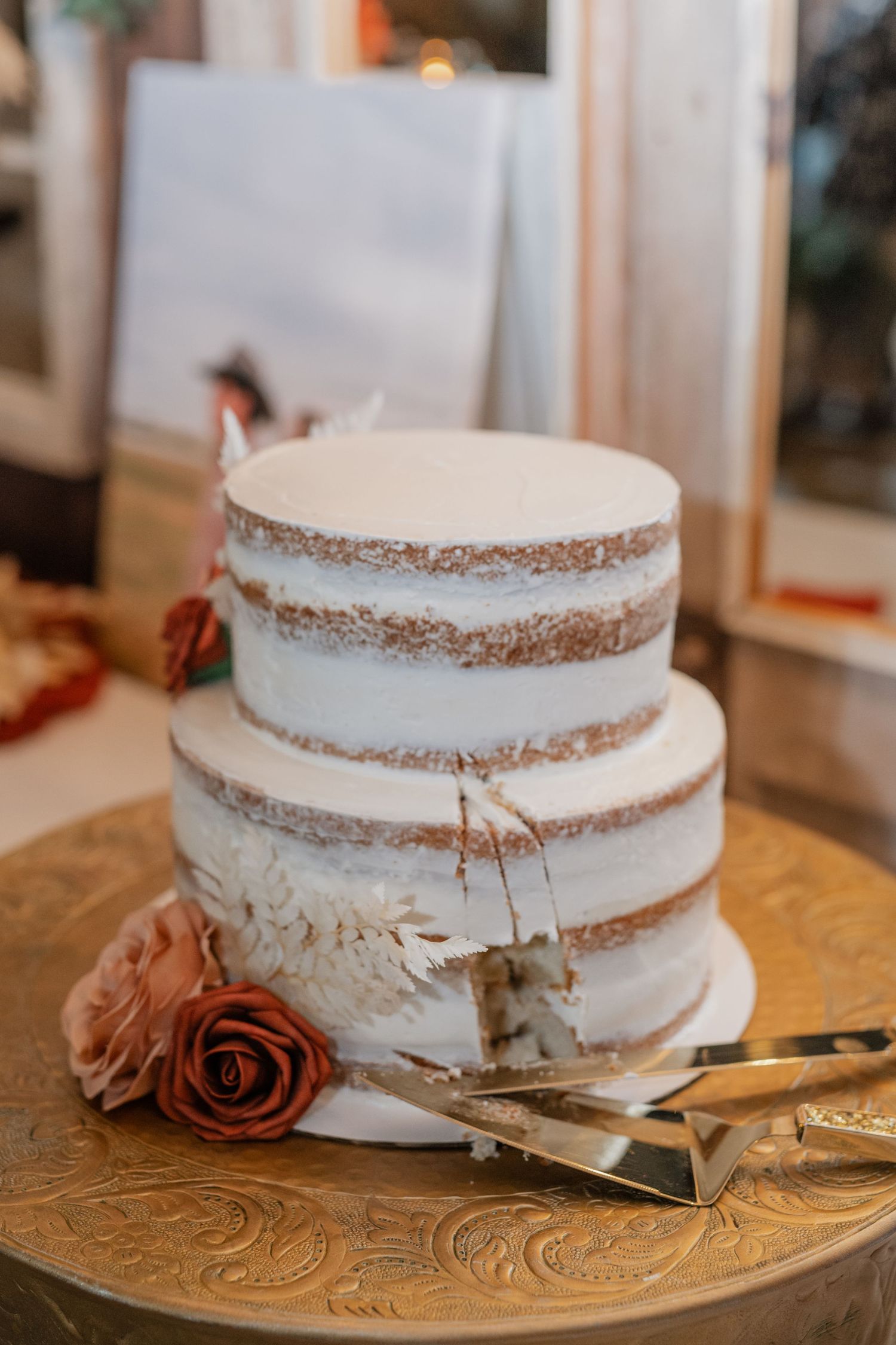 Elegant two-tier semi-naked wedding cake with white frosting and a dusty rose flower on a gold patterned cake stand.