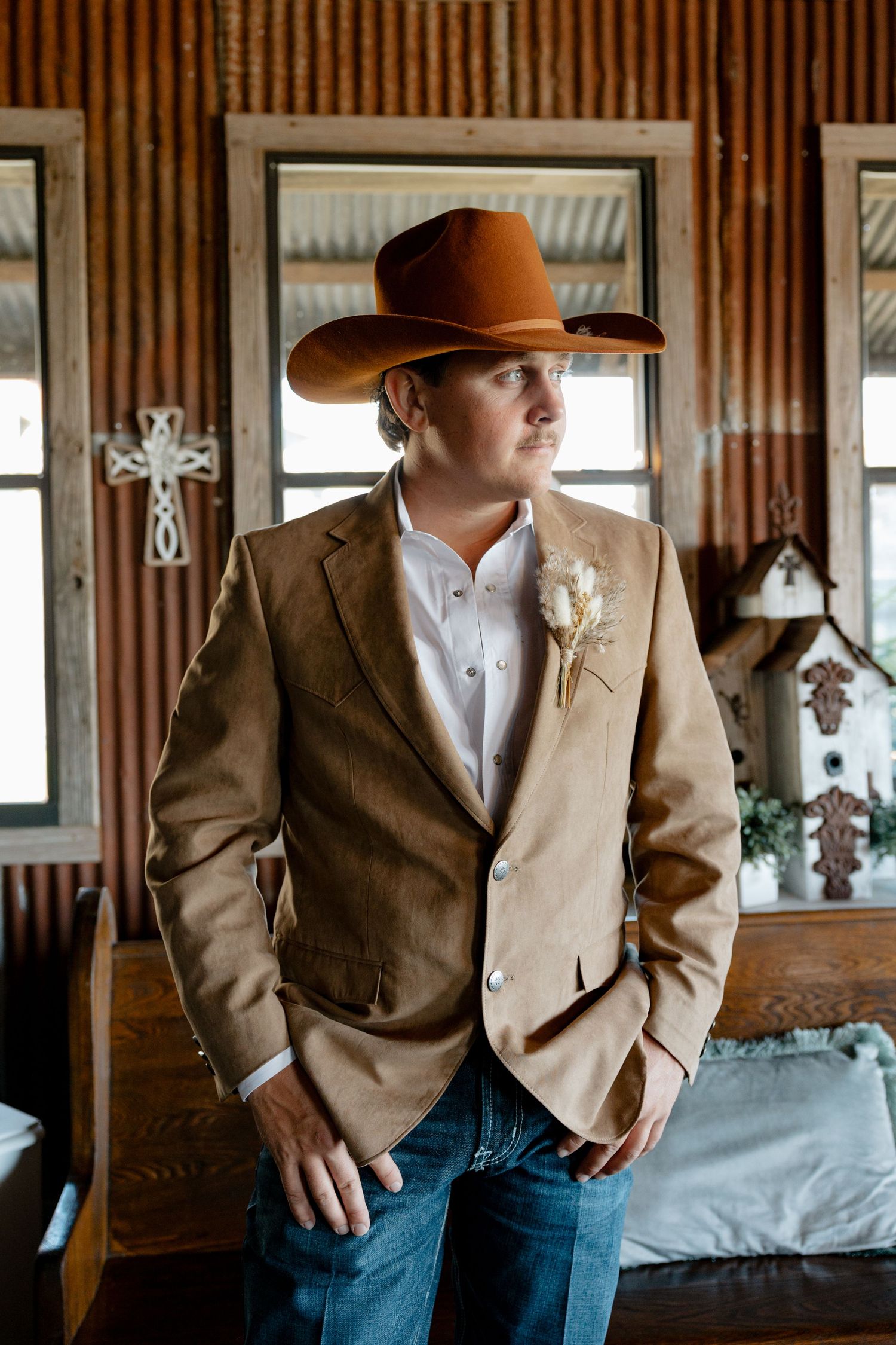Groom in brown suit jacket and cowboy hat standing in rustic wooden interior.