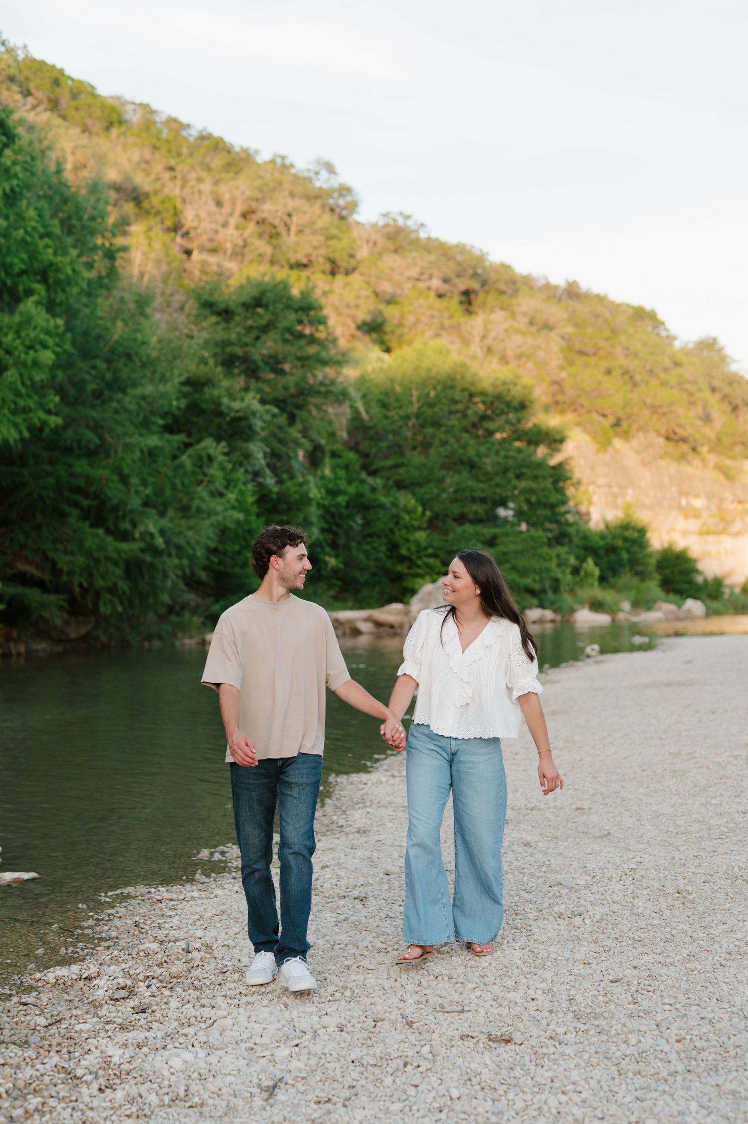 A couple walks hand in hand along a rocky riverbank path surrounded by green trees and hillside at sunset.