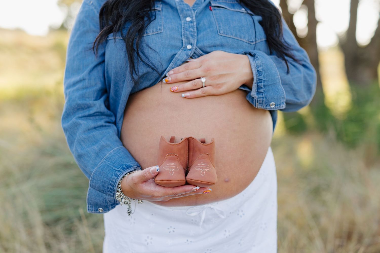 A pregnant woman cradles her baby bump while wearing a denim jacket in a natural outdoor setting.