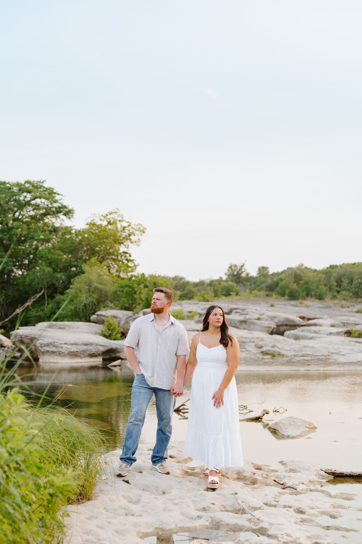 Couple explores and poses on rocky terrain near a peaceful river during sunset.