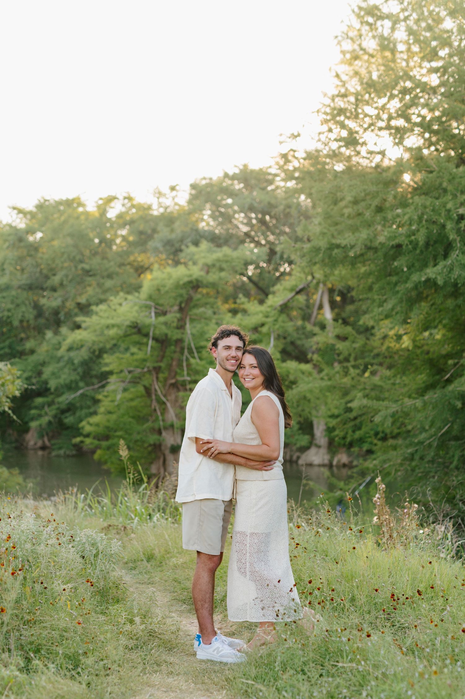 A romantic couple poses together in a sunlit meadow with wildflowers and green trees.