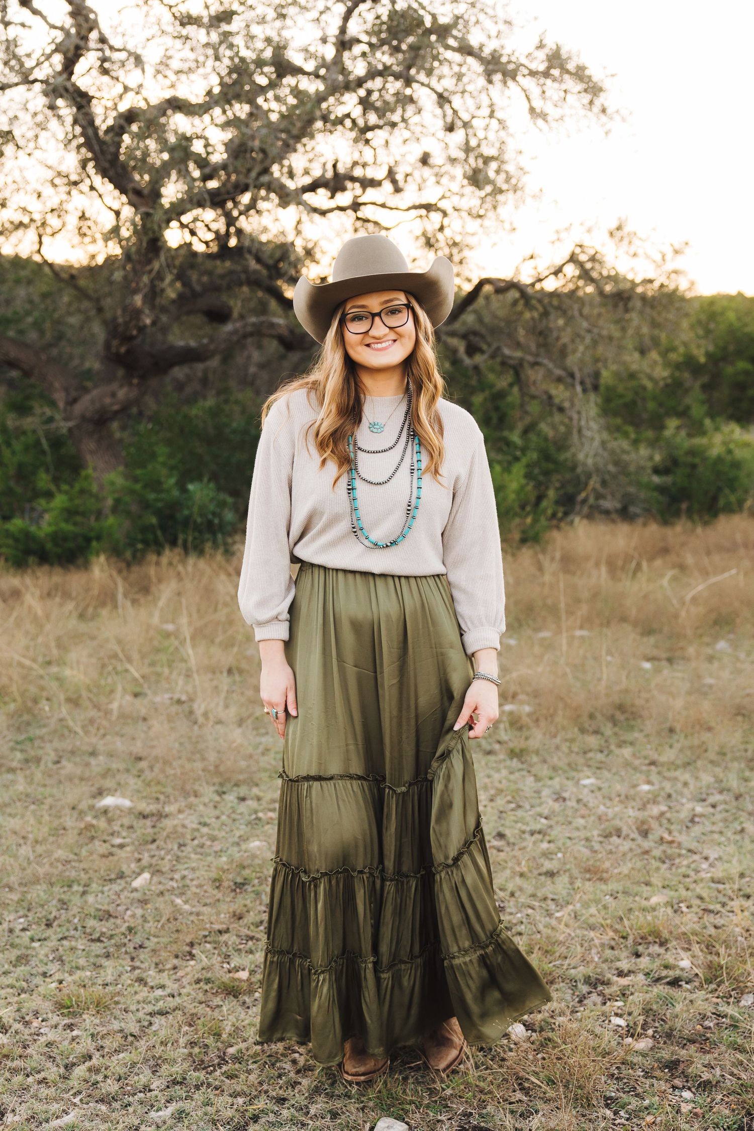 A western-style outfit featuring a tiered olive green maxi dress, white blouse, turquoise necklace and brown cowboy hat.