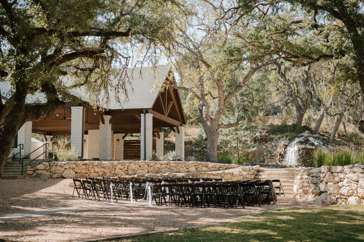 Rustic outdoor wedding venue with wooden chairs arranged in rows facing a small chapel under sprawling oak trees.