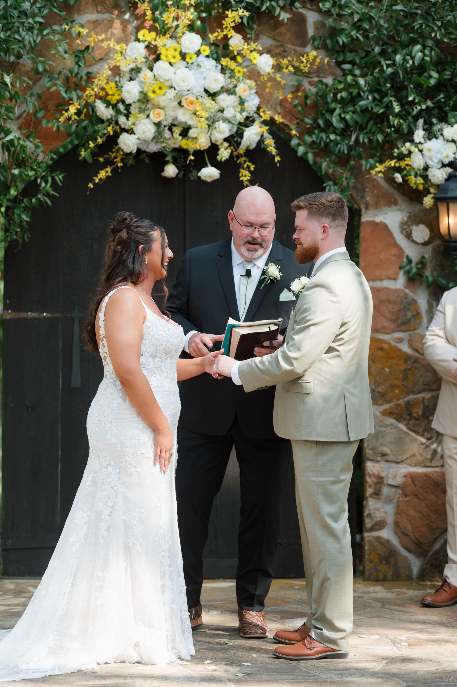 Bride and groom exchange vows with officiant under floral arch at outdoor stone venue.