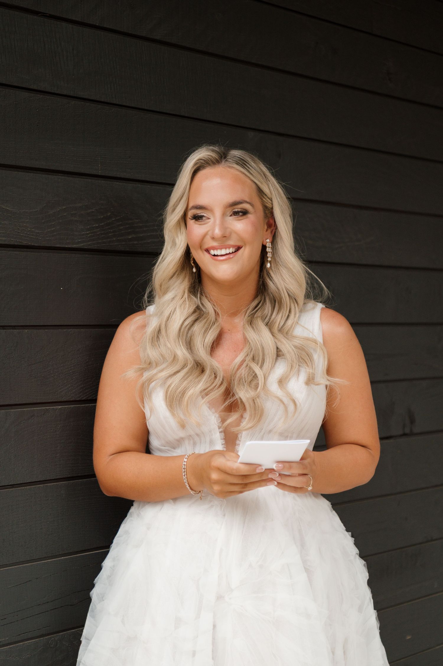 A smiling blonde woman wearing a white dress stands in front of a wooden wall.
