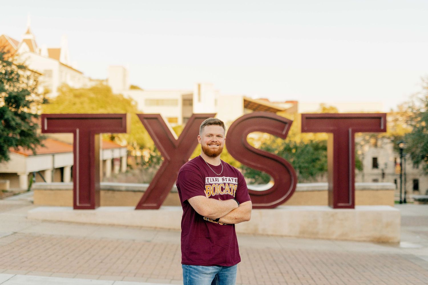 Graduate poses in front of TXST letters on Texas State University's campus during sunset wearing a Texas State University t-shirt.