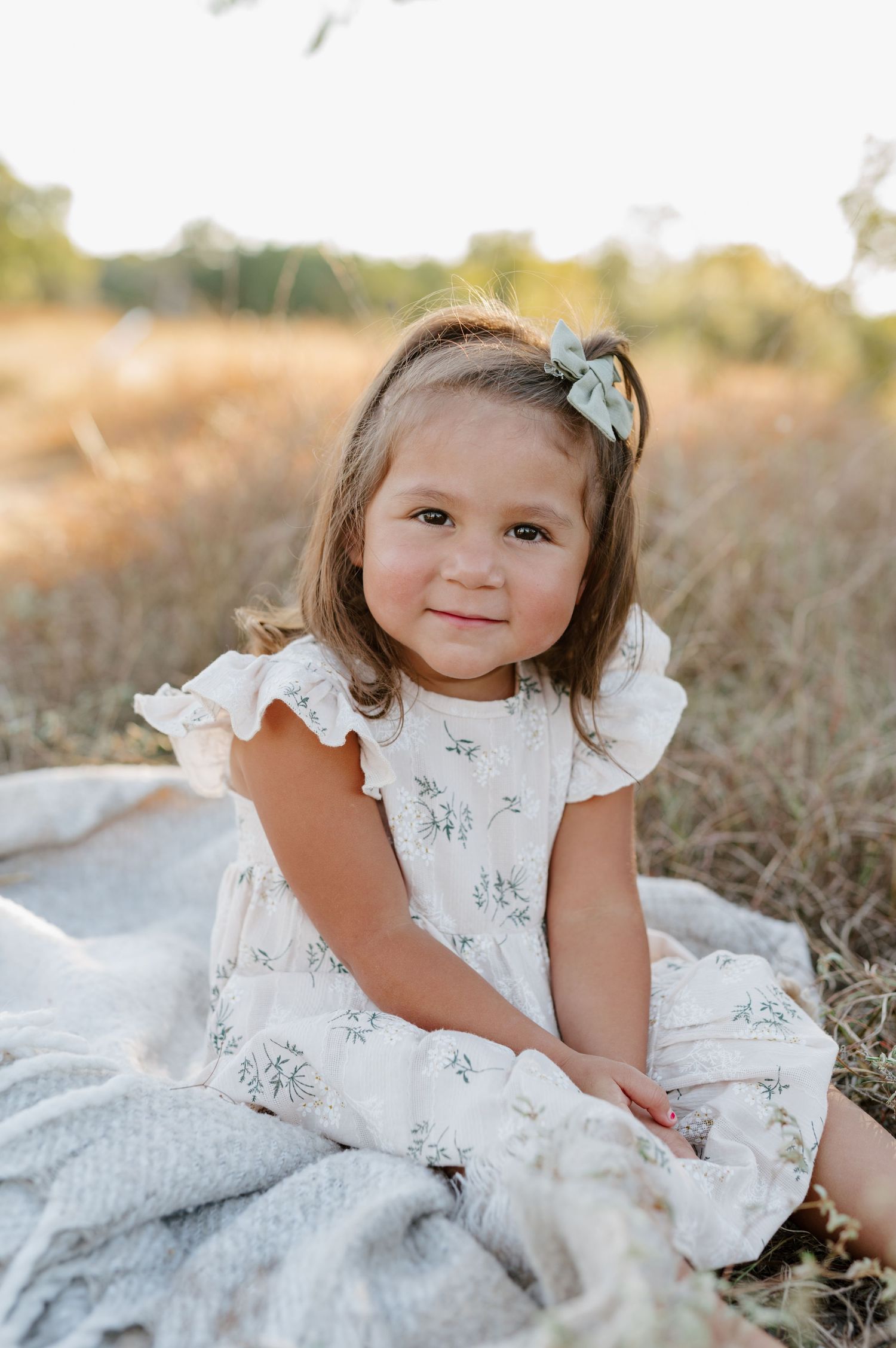 A small child in a white lace dress poses gracefully on a blanket in a sunny outdoor field.