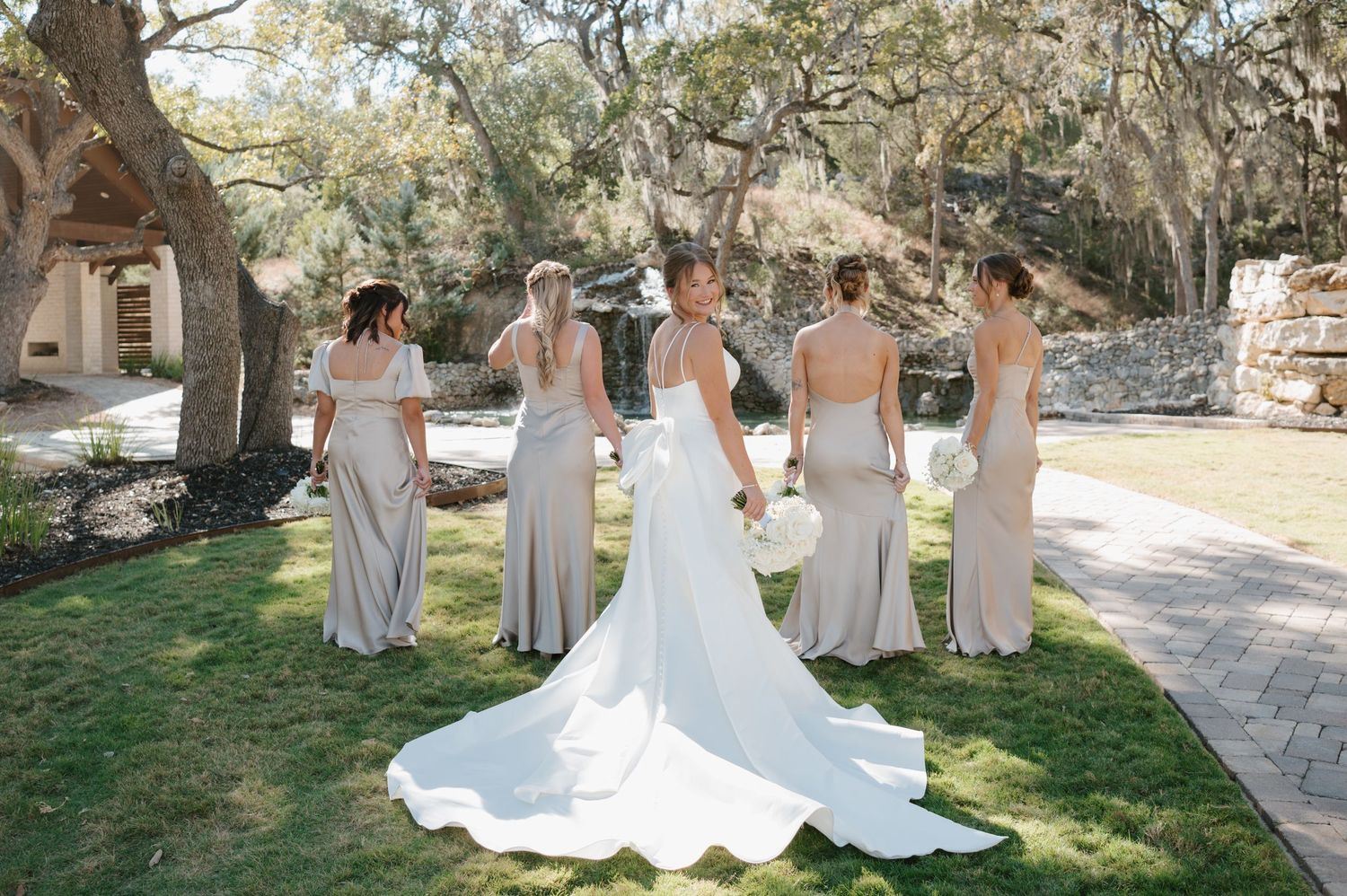 A wedding party in neutral colored dresses poses together outdoors under large oak trees on a sunny day.