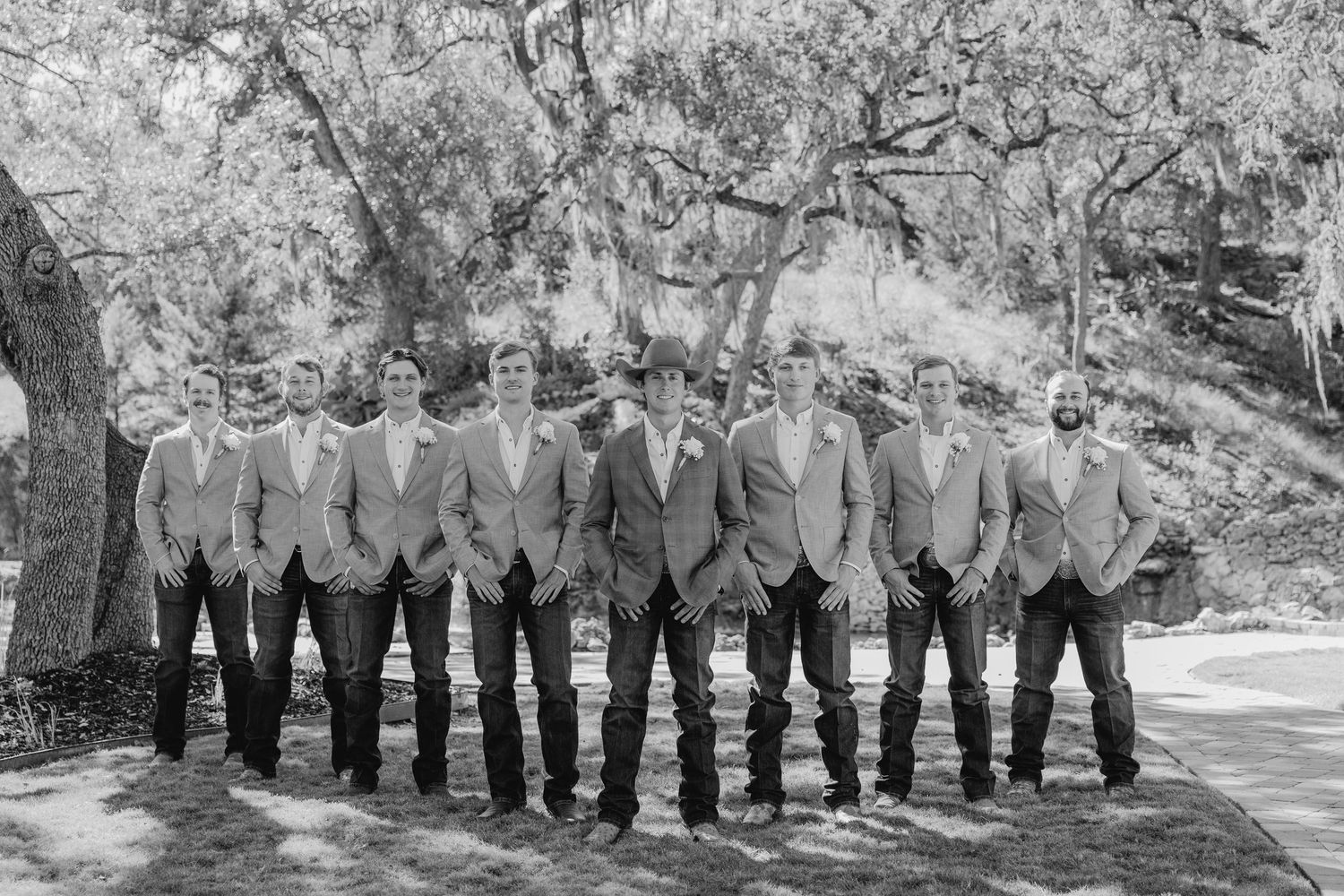 A line of groomsmen in formal attire standing outdoors against a natural backdrop.