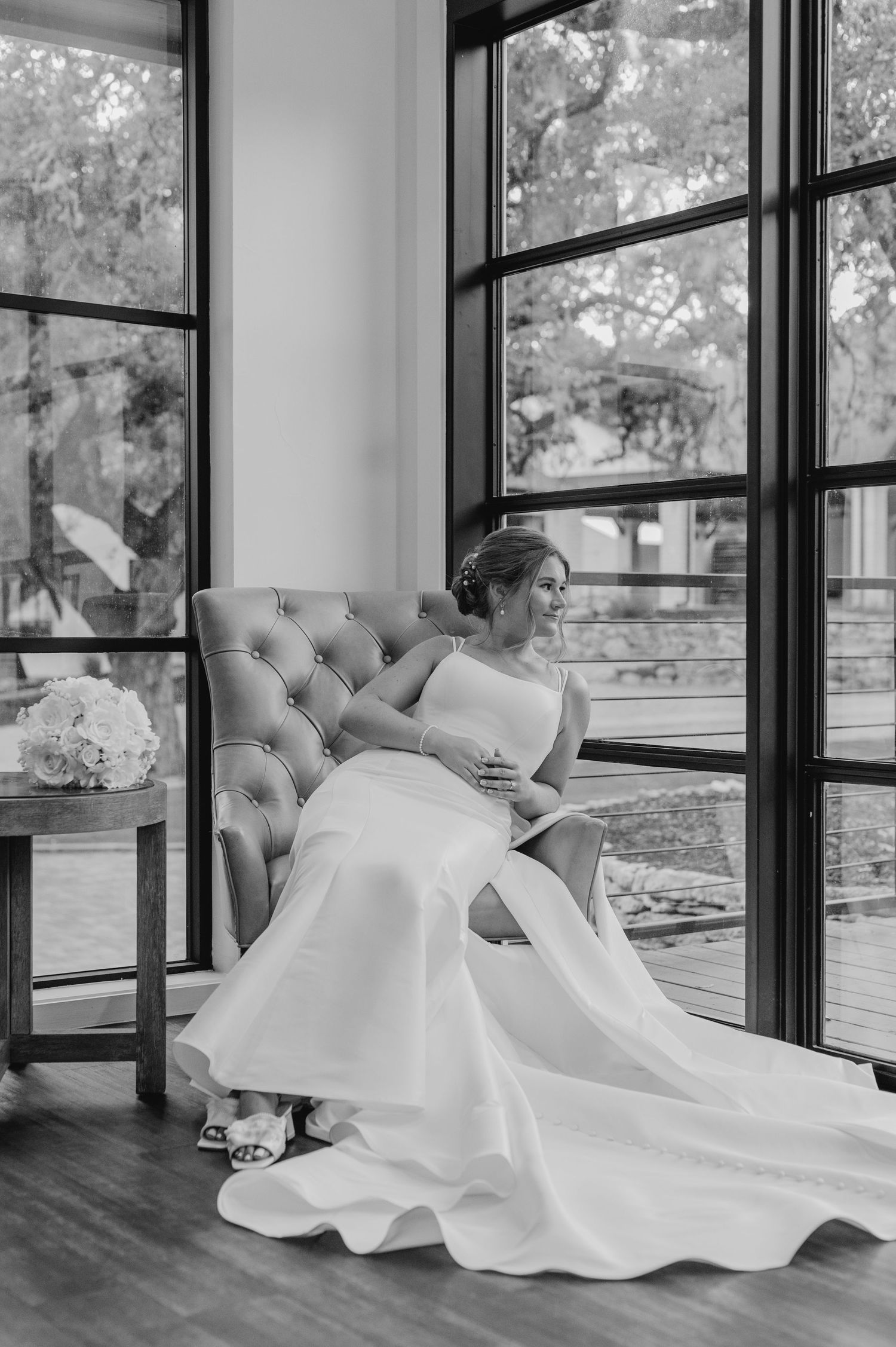 Wedding dress elegantly draped across grey tufted chaise lounge near large windows.