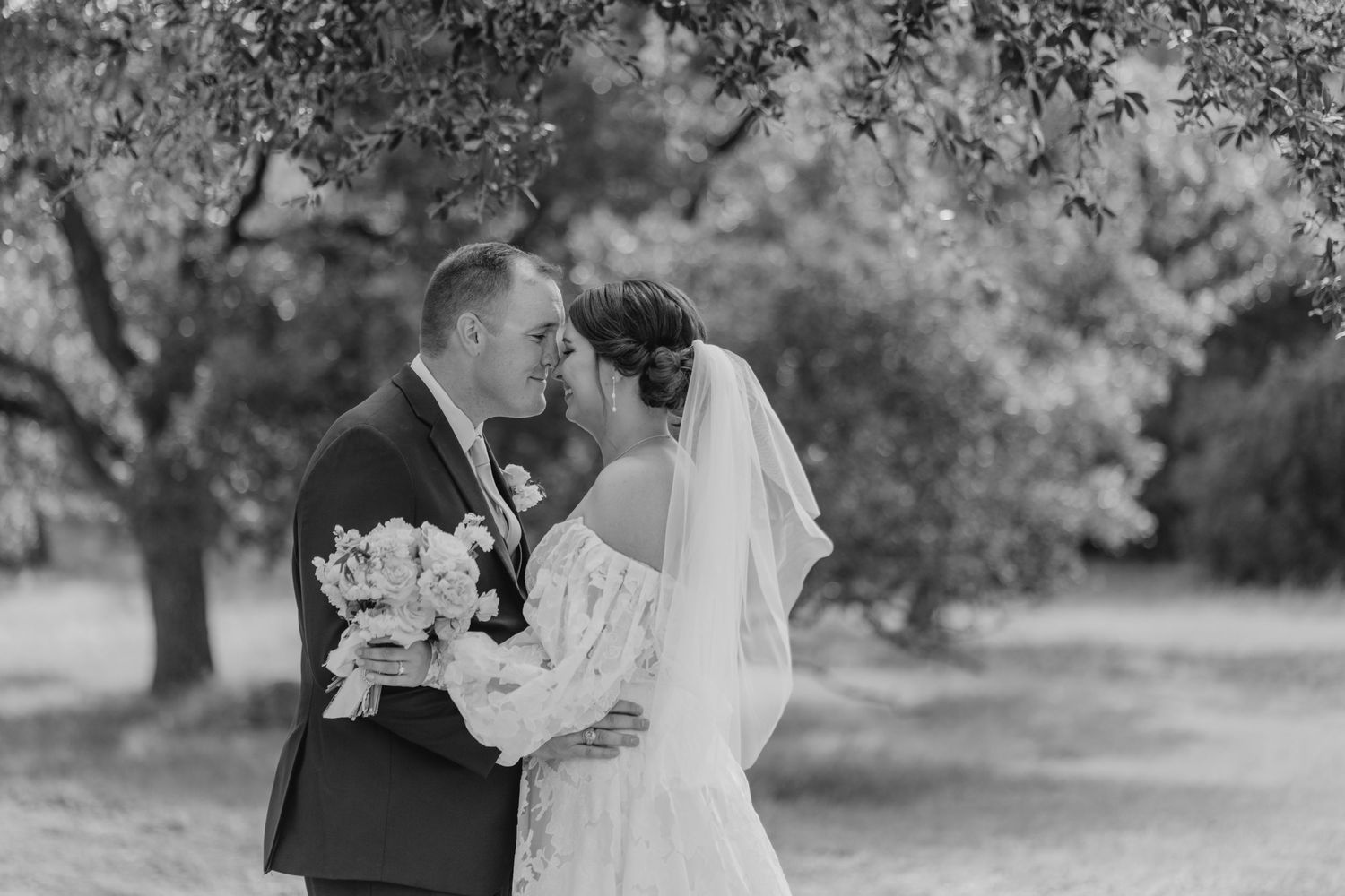 A bride and groom embrace in a garden setting, surrounded by trees and foliage.