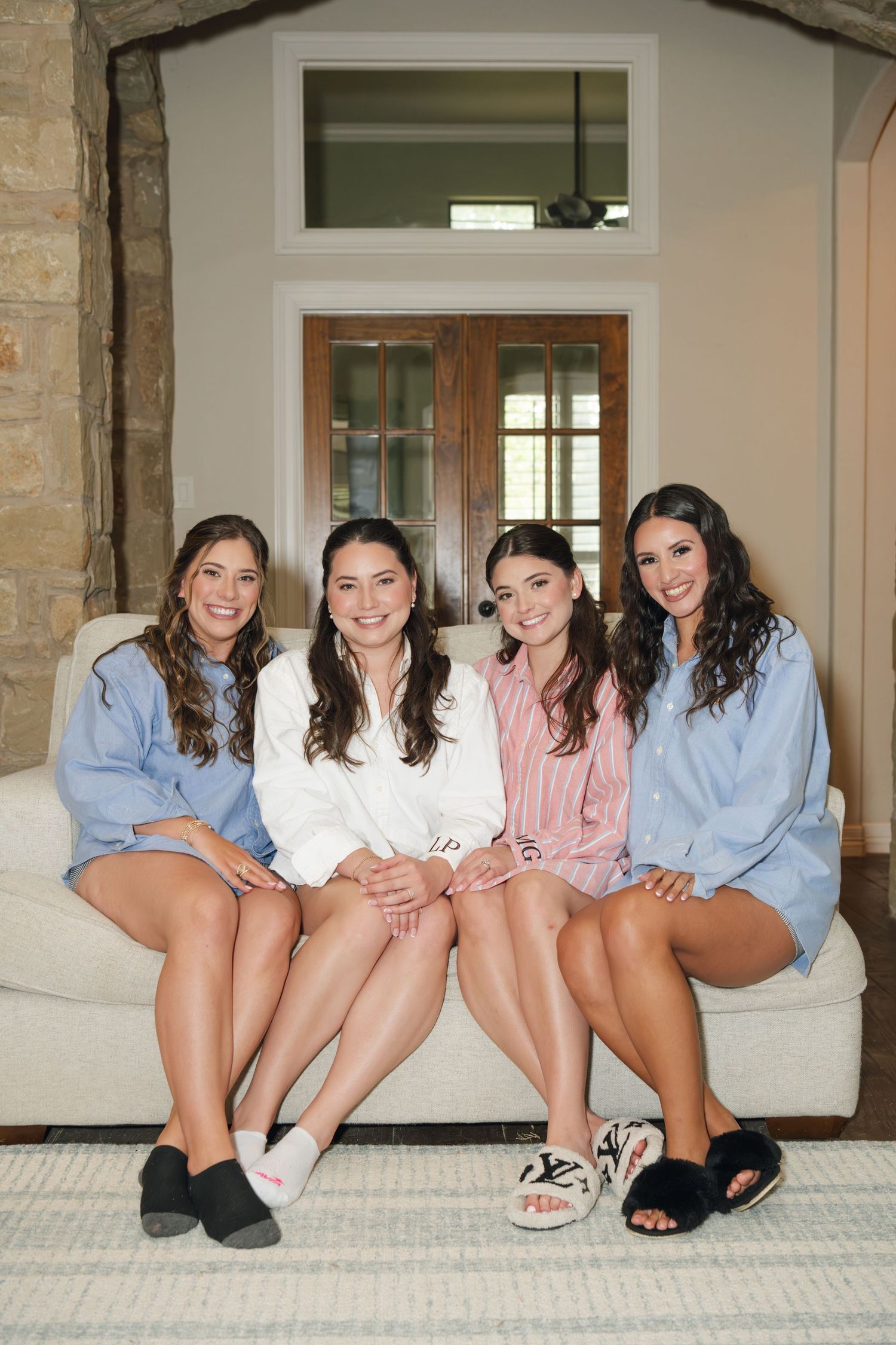 Four bridesmaids in matching robes and slippers sitting together on a white couch smiling before a wedding.