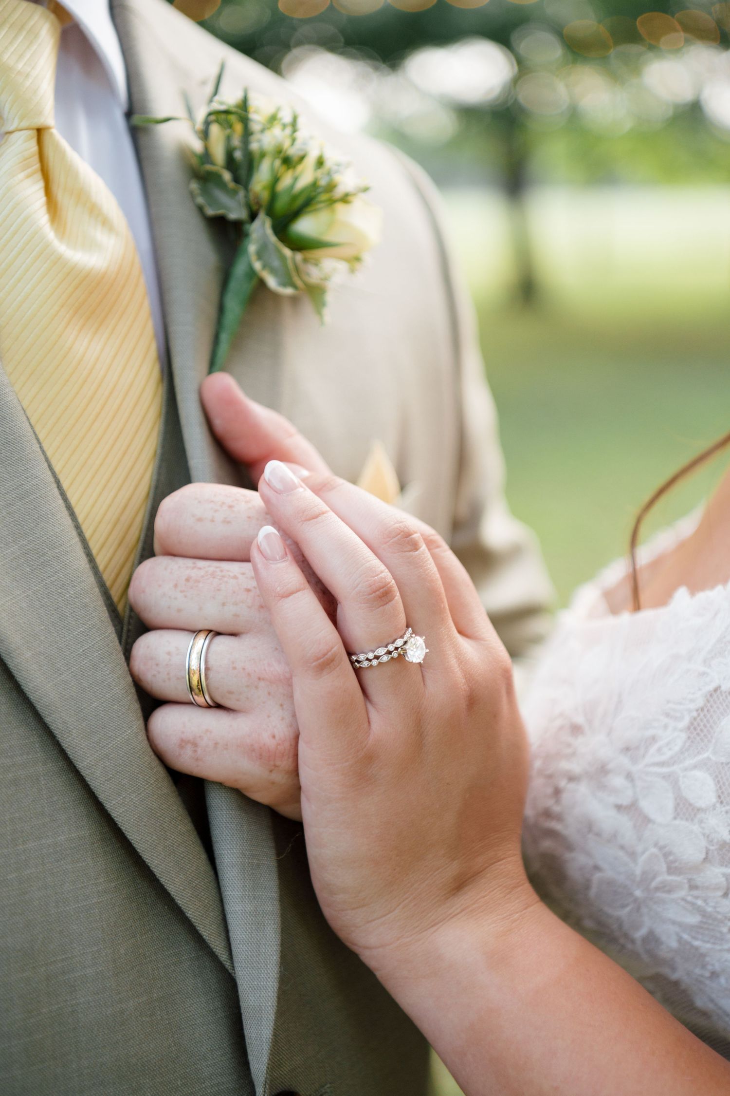 Close-up of bride and groom hands showing diamond engagement ring and wedding bands at outdoor ceremony.