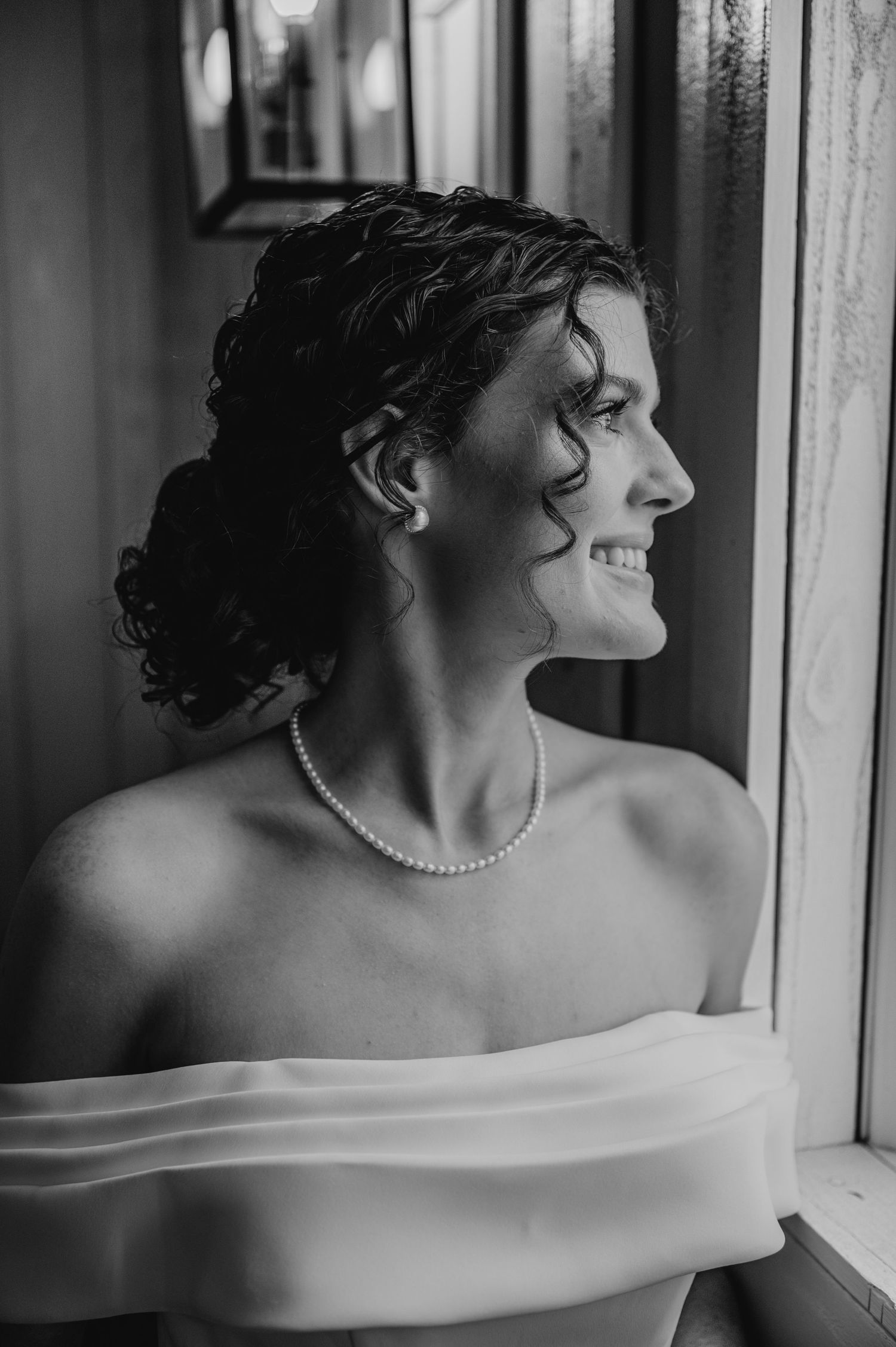 Black and white portrait of a bride in an off-shoulder dress with pearl necklace gazing out a window.