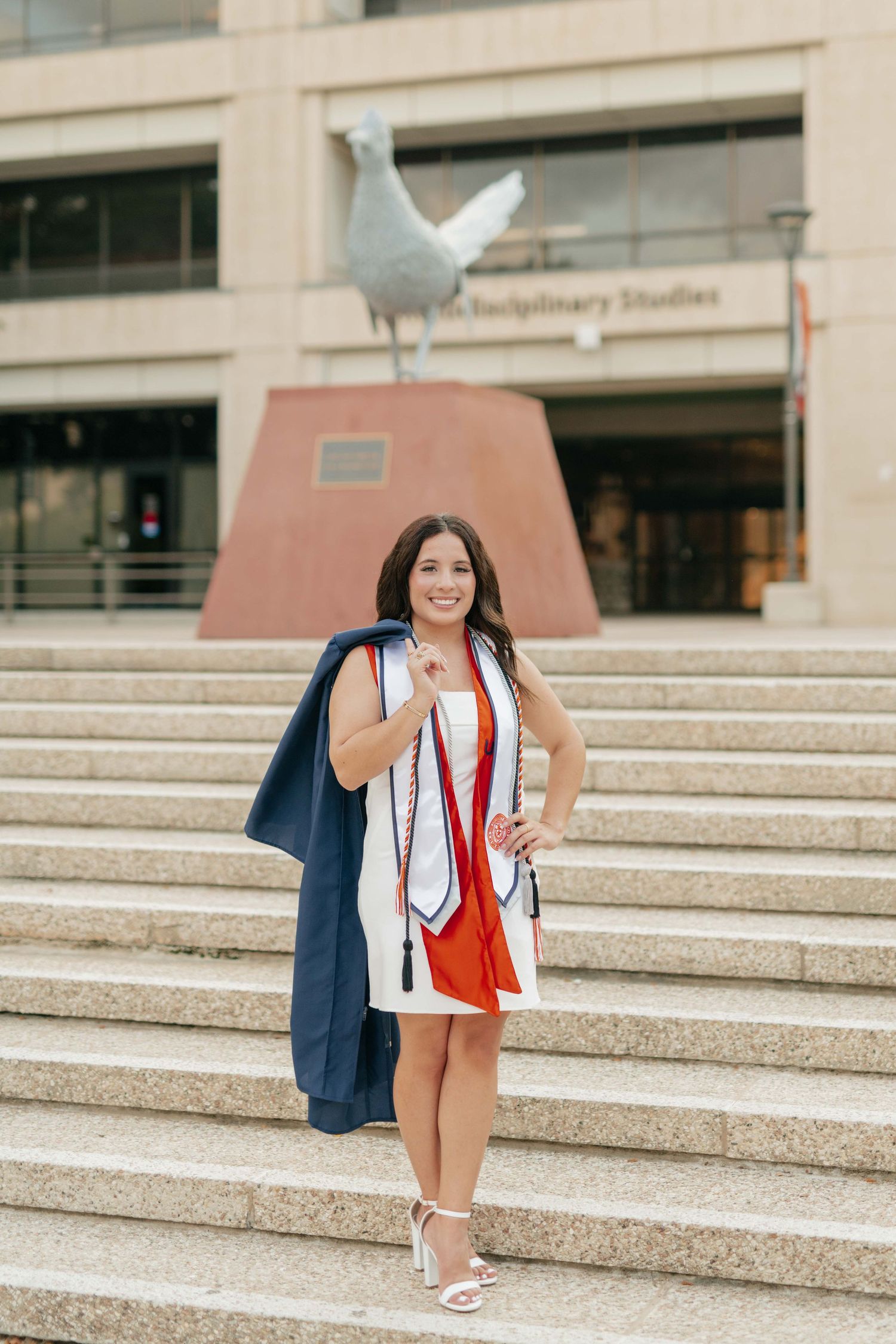 Graduate in white dress holding diploma on campus steps with seagull sculpture.