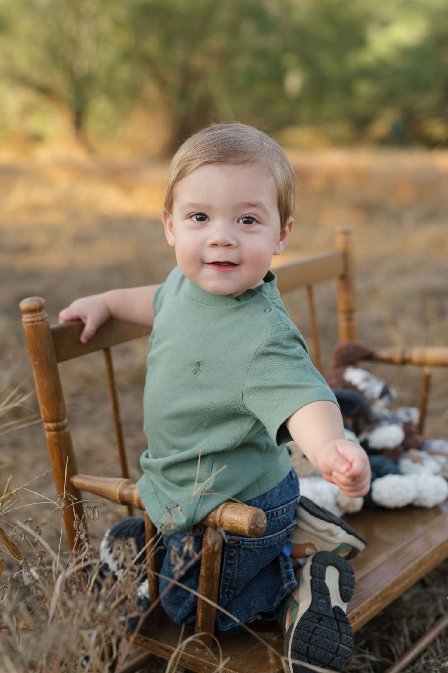A small child sits on a wooden bench outdoors wearing a green shirt and jeans among dried grass and cotton plants.