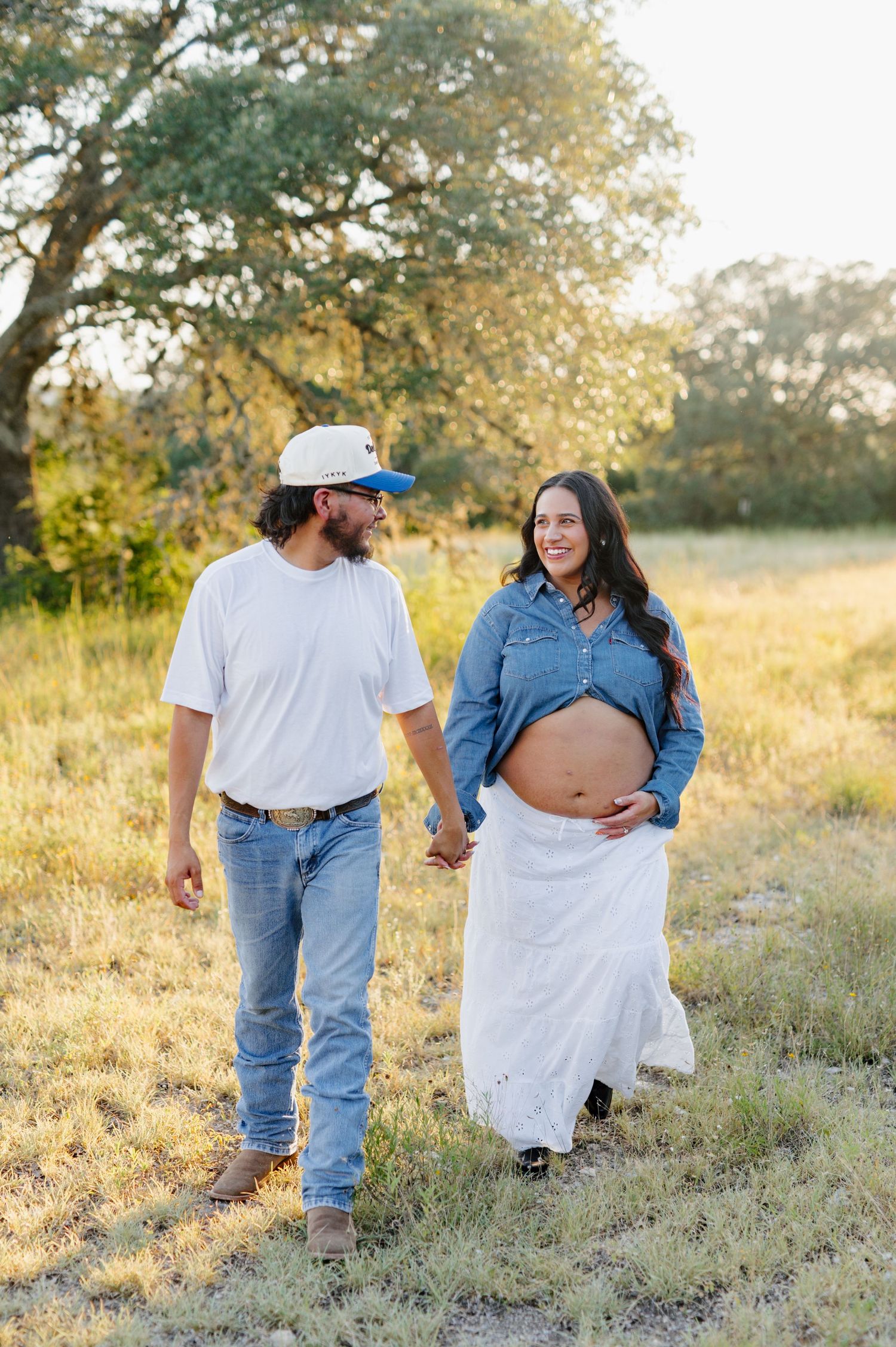A couple walks through a golden field during sunset for their maternity photoshoot.