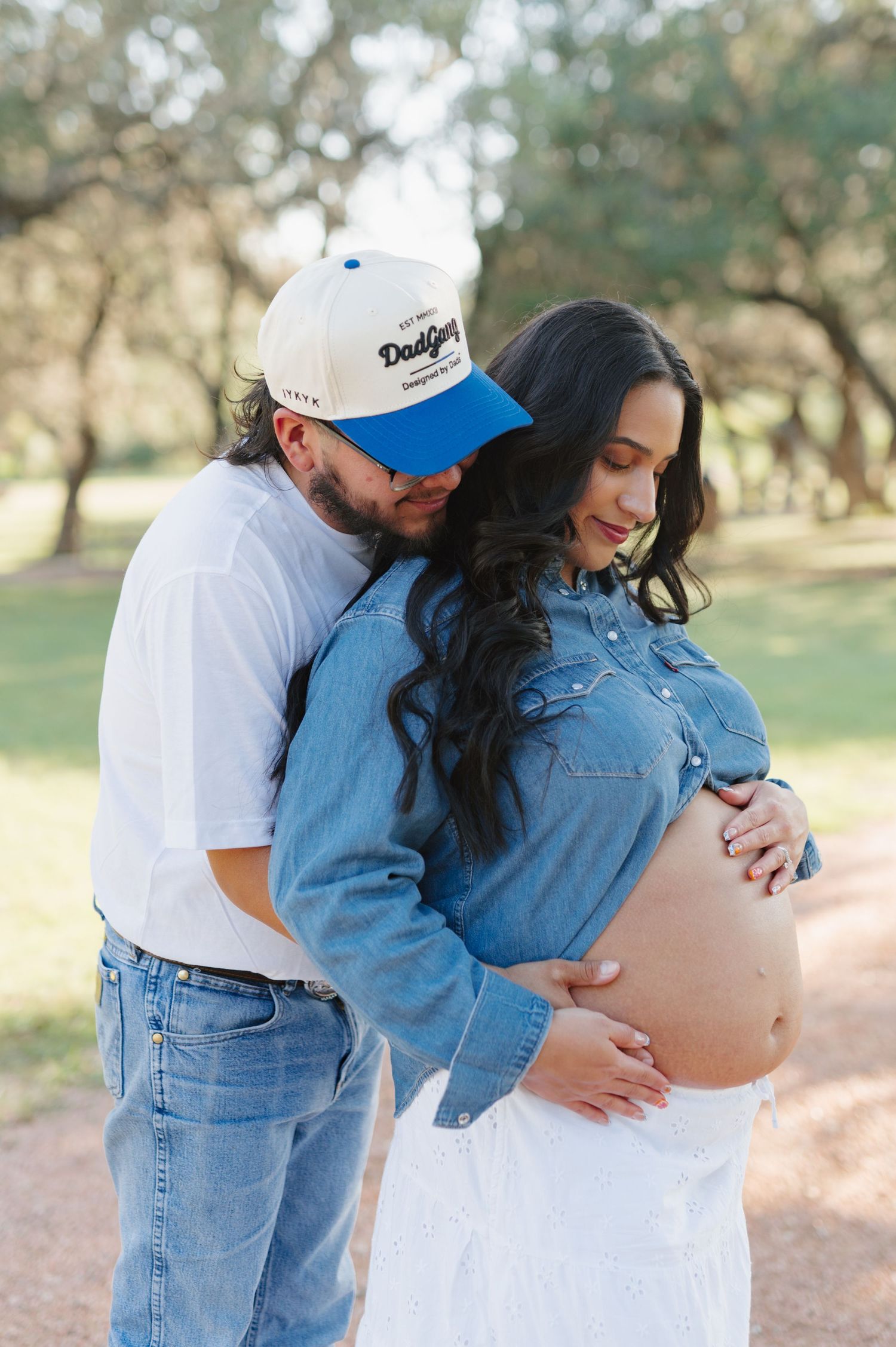 Expectant couple embraces while standing on a rustic path among trees during a sunset photoshoot.