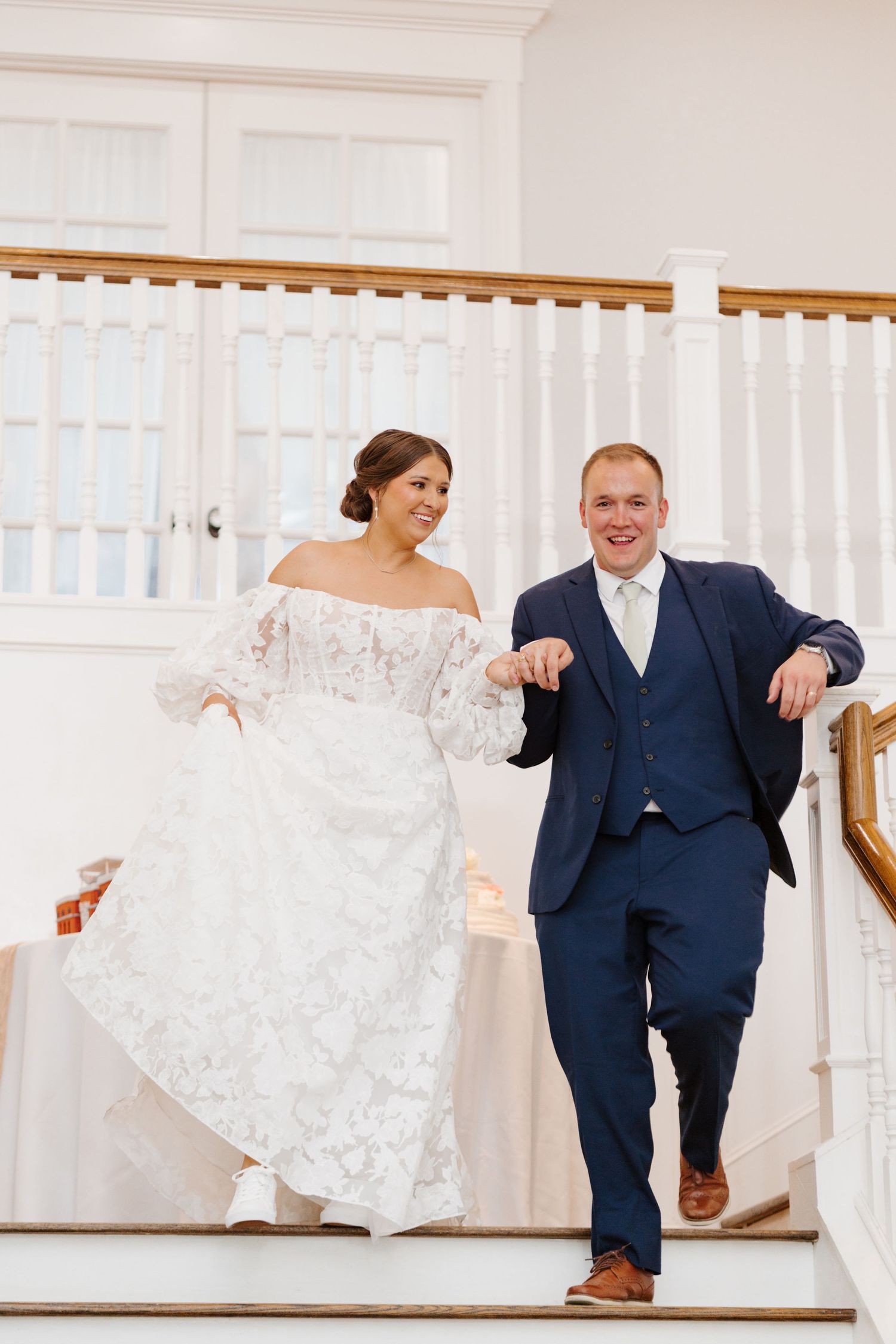 A couple runs down an elegant white staircase while holding the bride's lace dress train.