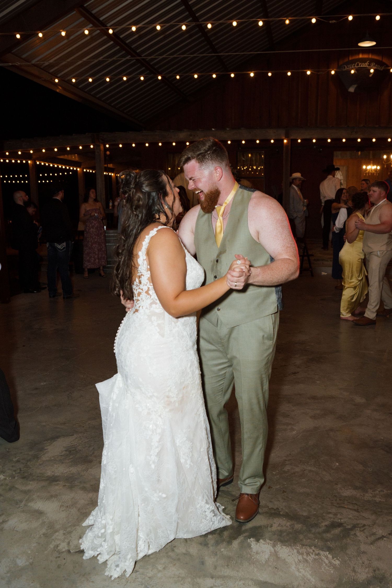 Bride and groom share a romantic dance under string lights at a rustic barn wedding reception.