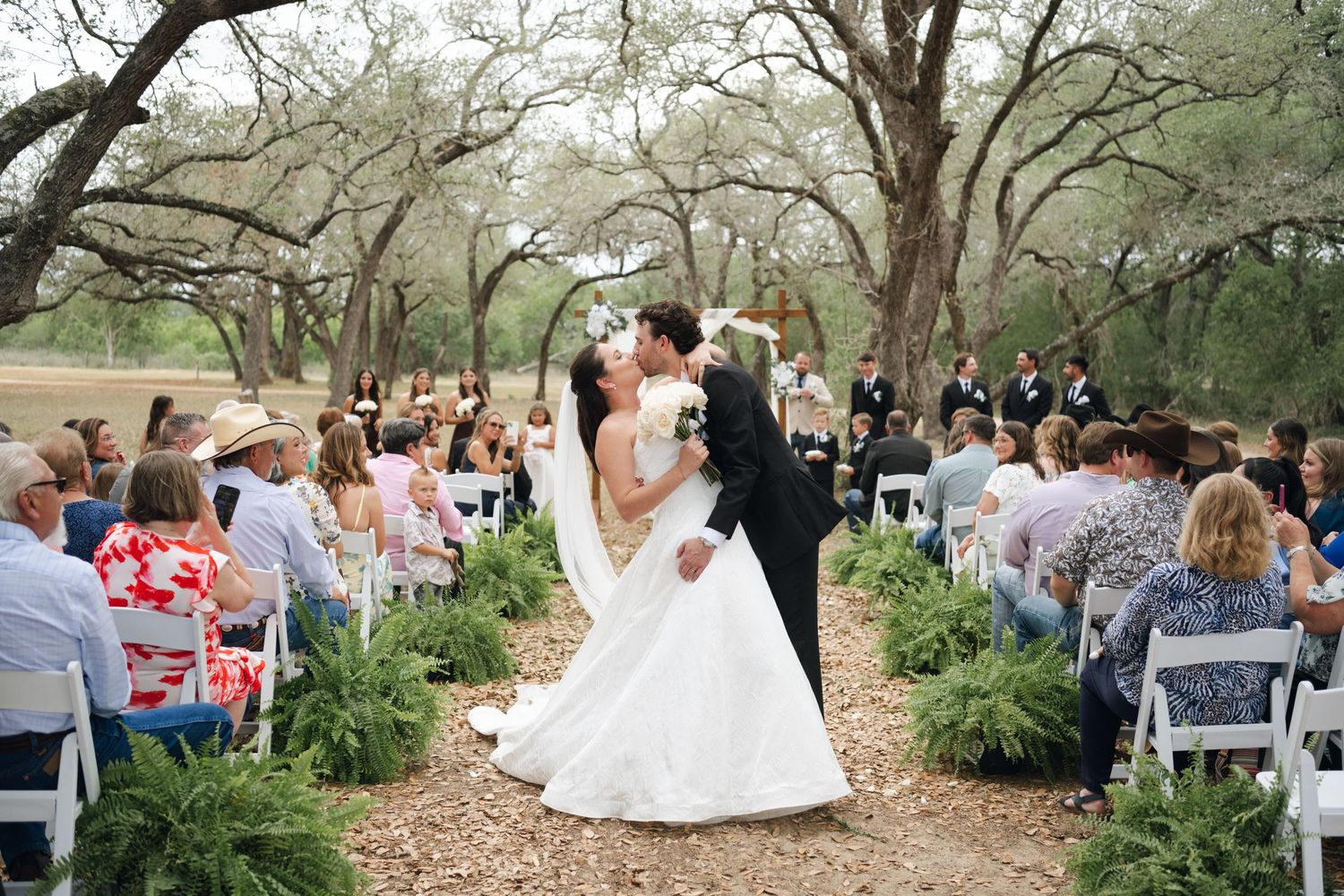 Newlyweds share first kiss at outdoor wedding ceremony aisle lined with ferns under oak trees.