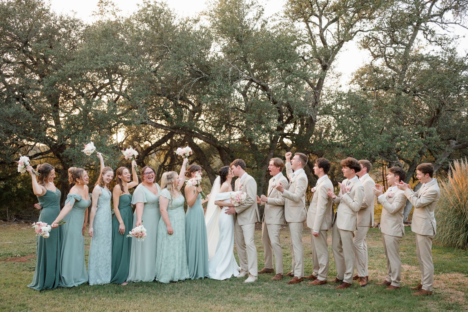 Wedding party in light blue dresses and tan suits poses outdoors under trees at sunset.
