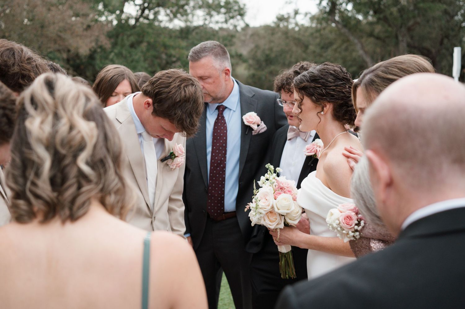 A group gathers at an outdoor wedding ceremony with formal attire and white floral bouquets.