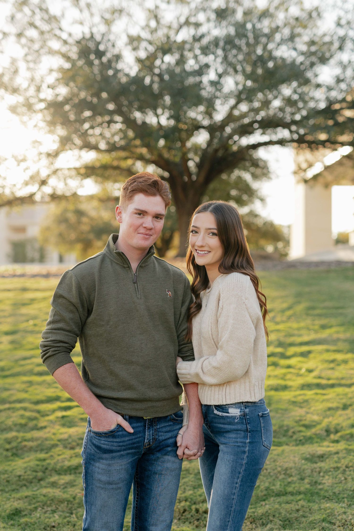 A couple in casual sweaters pose together on a grassy lawn at sunset.