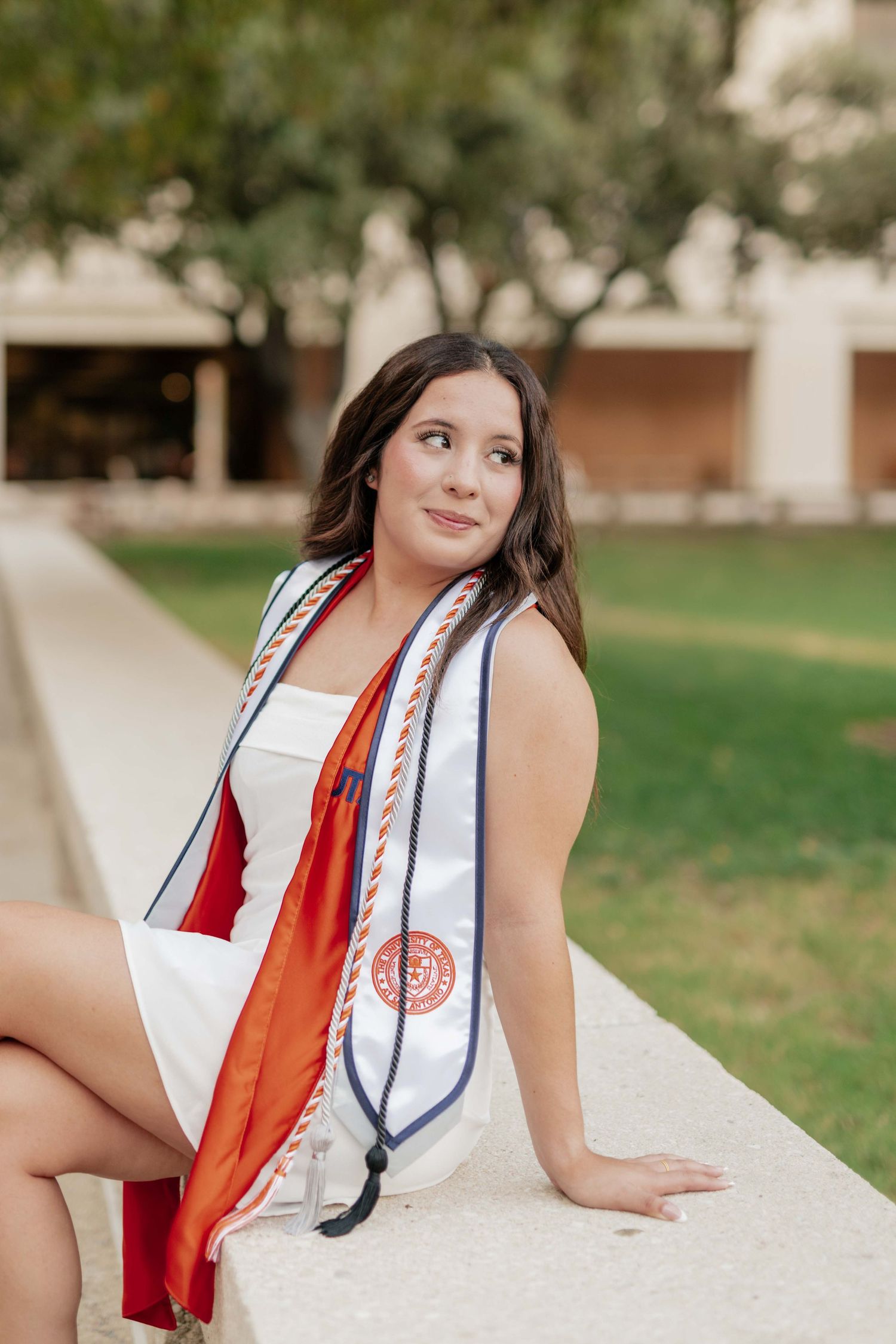 Graduate in white dress poses on campus wall with decorative stole.