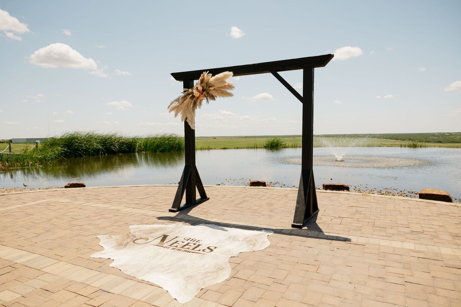 A wooden wedding arch decorated with burlap stands on a lakeside beach with a white carpet runner on the sand.