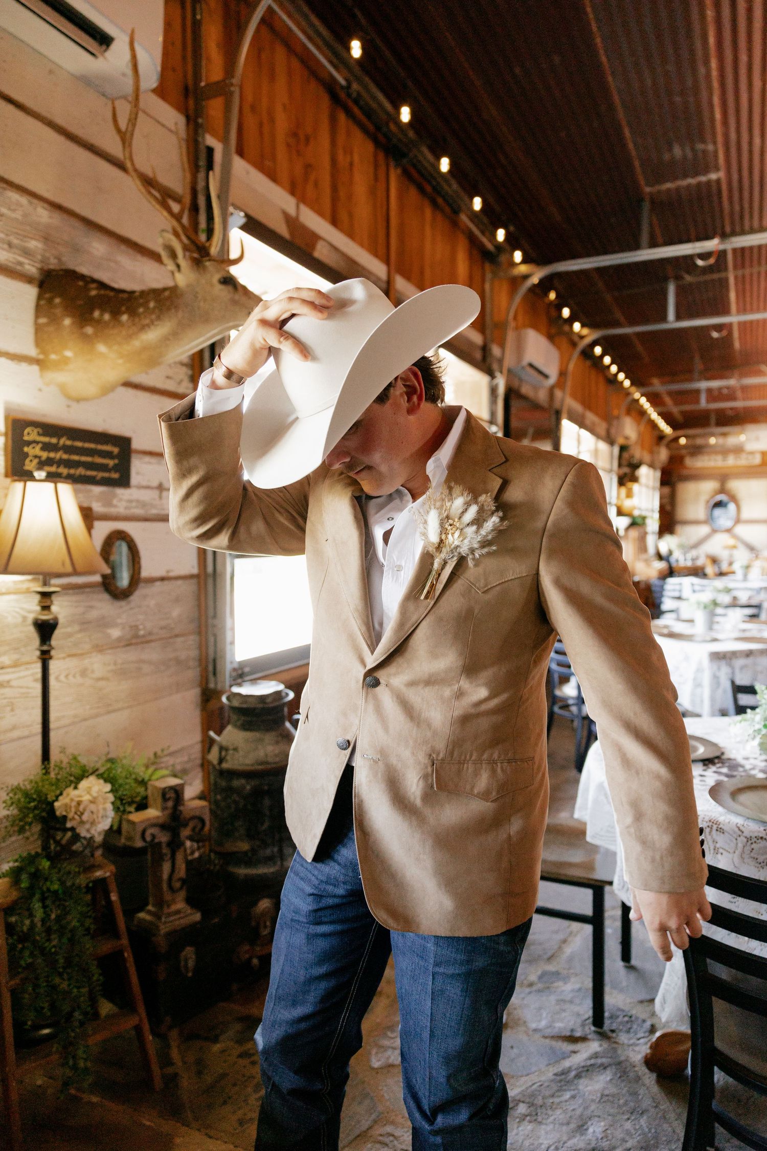 Country western styled groom in tan blazer and cowboy hat in rustic indoor setting.