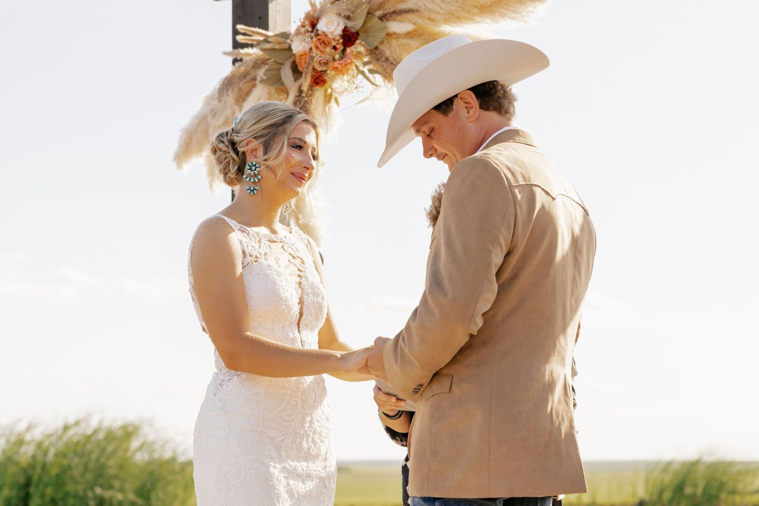 A couple holds hands during their western-themed outdoor wedding ceremony against a sunny field backdrop.