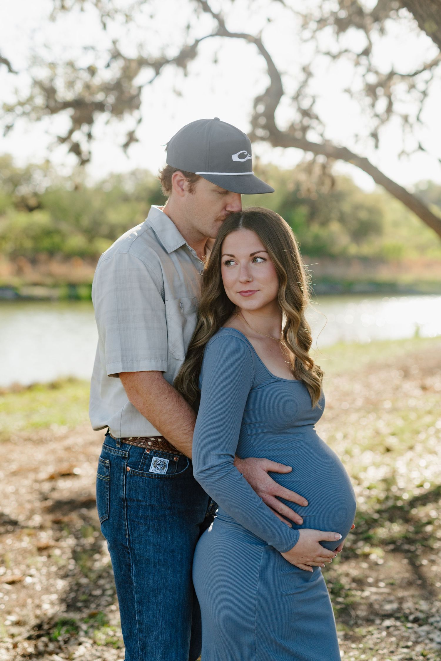 A couple shares a tender moment by a lake, with the expectant mother wearing a blue dress that highlights her baby bump.