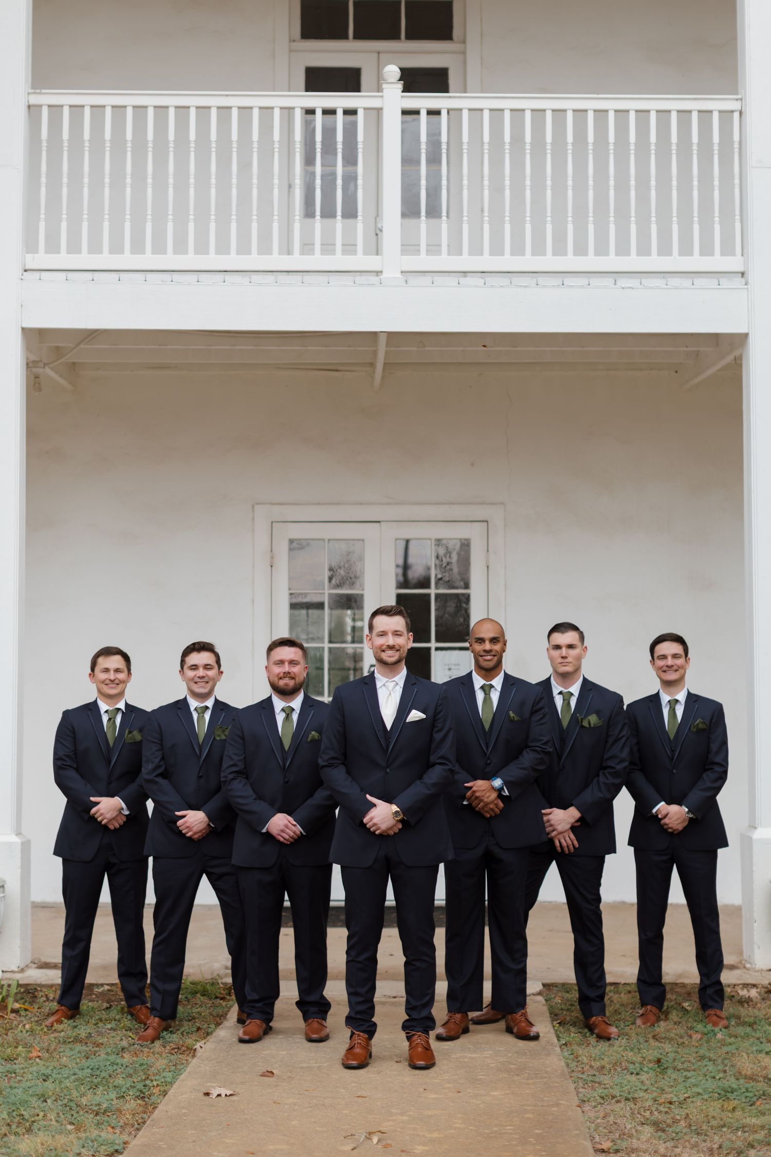 Groomsmen in black suits and brown shoes sitting on white porch railing.