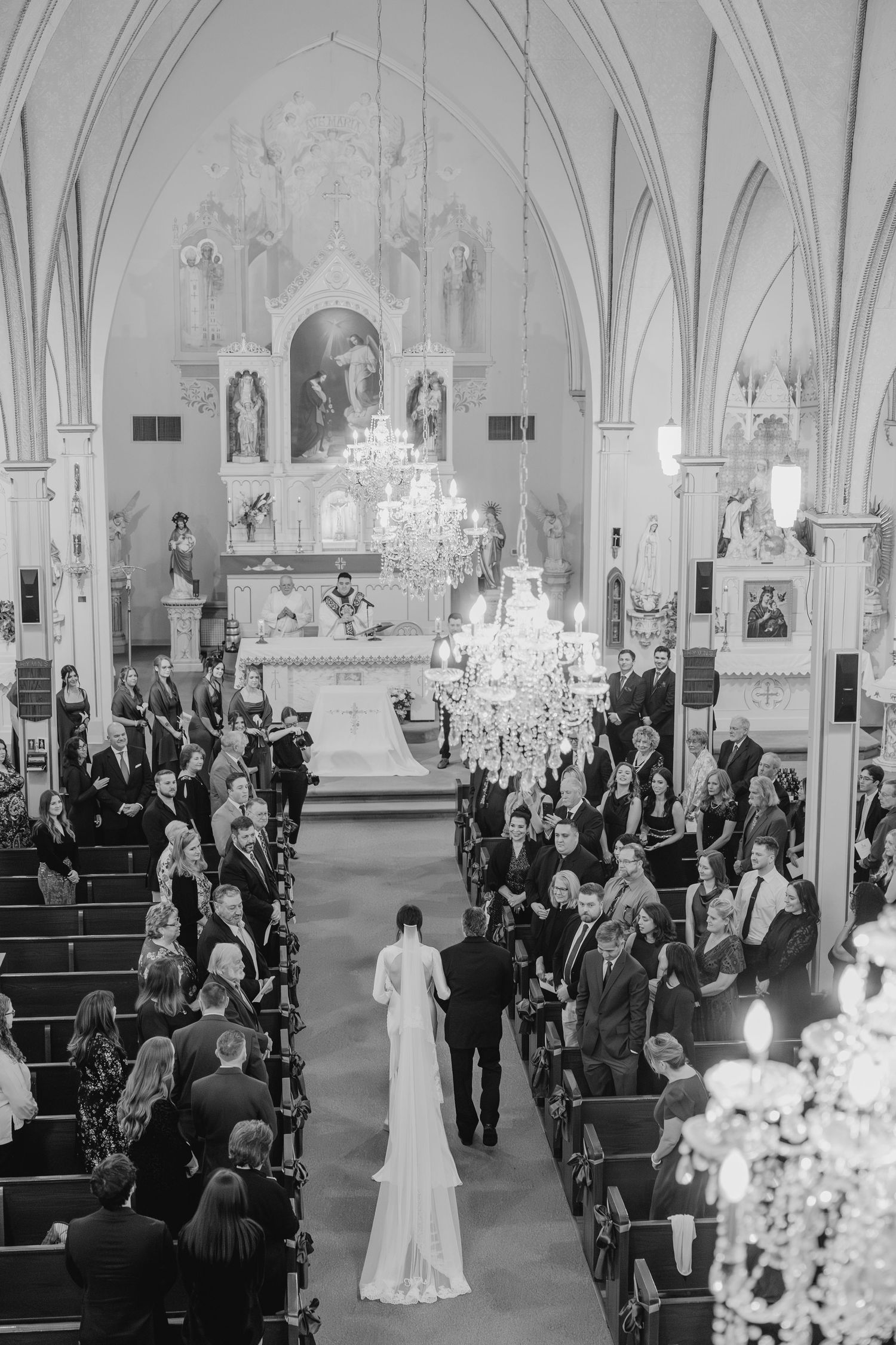 Wedding ceremony in a grand cathedral with ornate architecture, chandeliers, and guests seated in wooden pews.