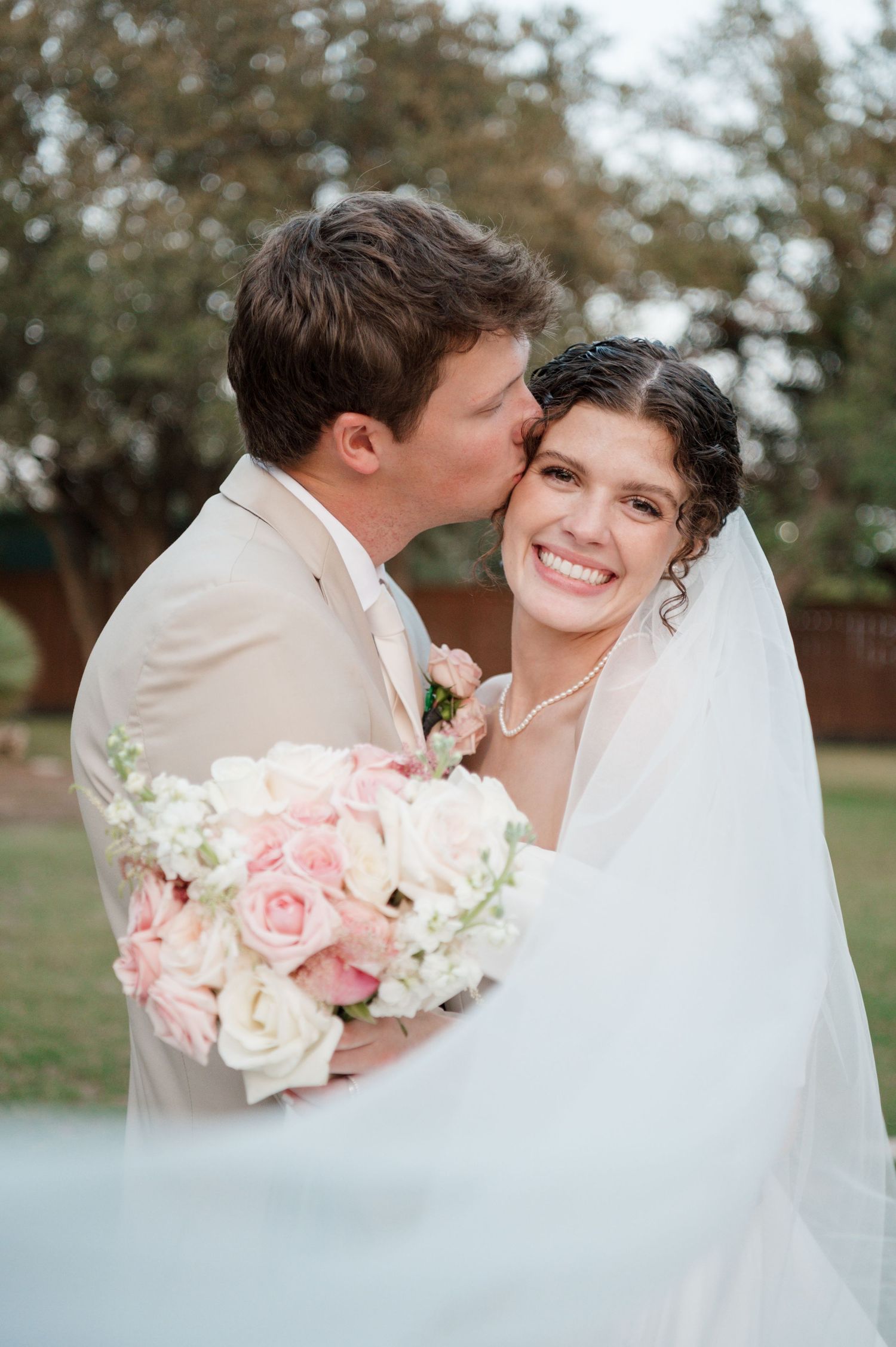 A romantic wedding couple shares tender moments in a garden setting with soft pink roses and white florals.