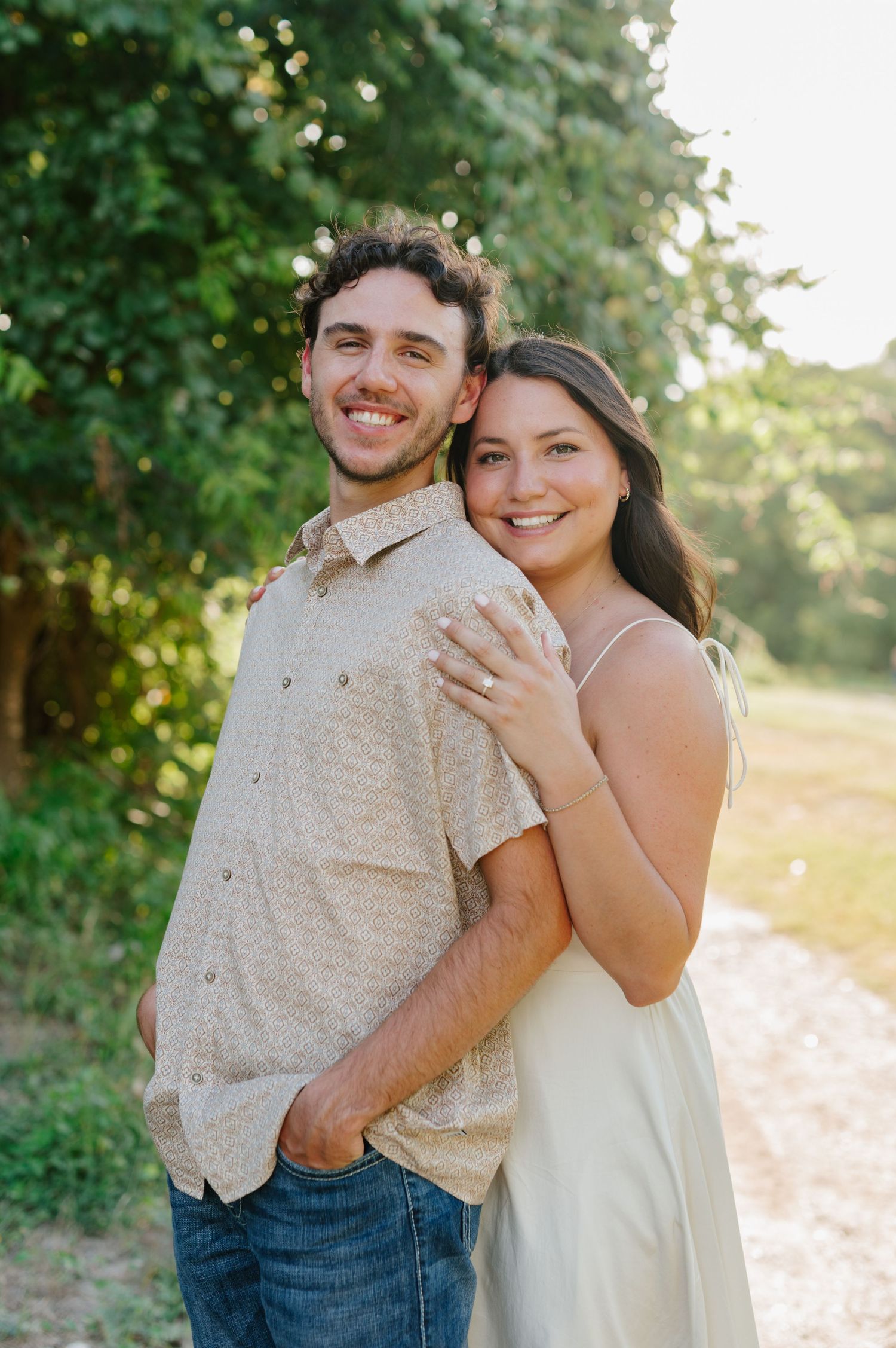 A couple embraces against a sunlit backdrop of green foliage in a romantic outdoor setting.